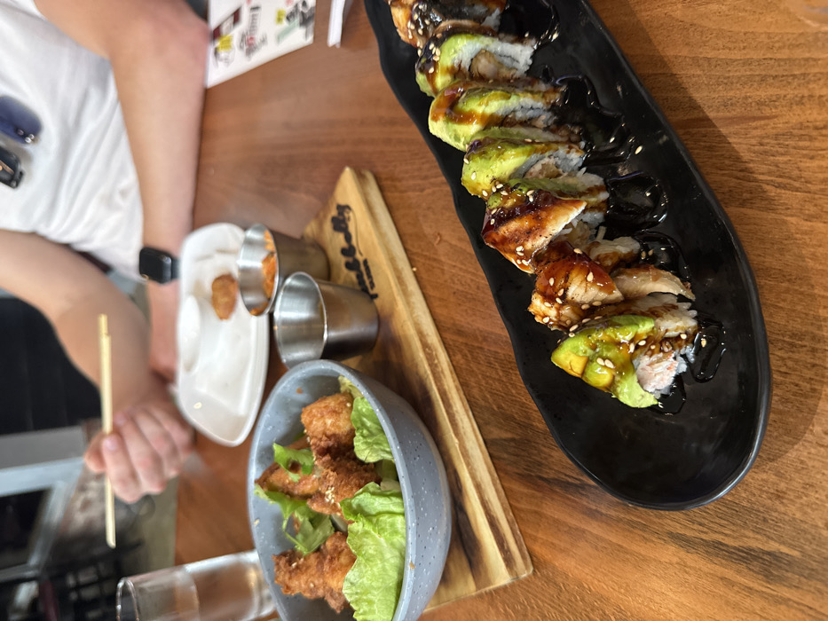 The scene depicts an indoor dining environment at Humbelly Burger & Sushi, located in Miami, Florida, United States of America. A wooden table serves as the primary surface. In the foreground, a black, oblong ceramic plate holds nine pieces of sushi, each topped with sliced avocado, a piece of glazed eel, a dark sauce drizzle, and white sesame seeds. To the mid-left, a grey speckled bowl contains several fried pieces of food, possibly chicken karaage, resting on green lettuce leaves and sprinkled with sesame seeds. Positioned centrally on a wooden serving board, which bears the partially visible brand name "humbelly," are two small stainless steel ramekins; one contains a reddish-orange condiment, and the other holds a dark liquid sauce. In the upper right, an individual is partially visible from the chest up, wearing a white short-sleeved shirt and an Apple Watch on their left wrist. Their right hand holds wooden chopsticks, positioned above a white rectangular plate that contains a single fried, golden-brown nugget. Dark sunglasses are clipped to the person's shirt collar.