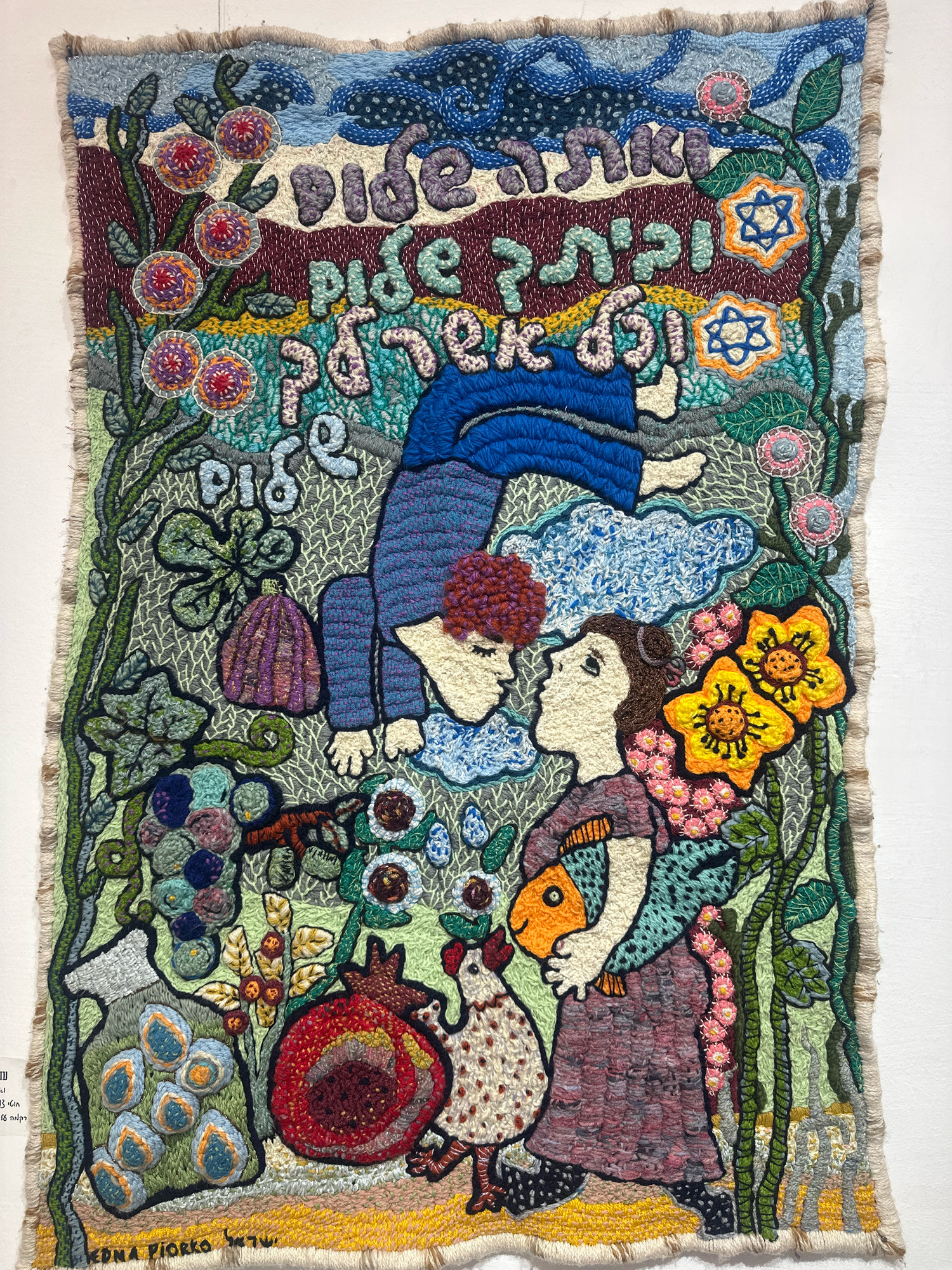 This image displays a colorful, textured tapestry or rug, likely an artwork, depicting a scene with stylized figures and symbolic elements. The tapestry is predominantly in shades of blue, green, purple, yellow, orange, and red, with intricate stitching and tufting that creates a rich, tactile surface.
At the top, Hebrew text is woven into the fabric. Below the text, two stylized figures are depicted. One figure, dressed in blue, is in a dynamic, almost acrobatic pose, with their body contorted as if suspended in the air or reaching for something. Their face is turned downwards towards another figure. This second figure, facing upwards, has brown hair and is looking towards the suspended figure. They are holding an orange fish in their arms and are accompanied by a white and brown speckled chicken at their feet. The figures are surrounded by a vibrant, nature-inspired environment. Lush green vines with leaves, clusters of grapes, and a large red pomegranate are visible, along with stylized flowers in various colors, including yellow and pink. Blue clouds are also integrated into the composition, particularly around the suspended figure. Two Star of David motifs are woven into the upper right portion of the tapestry. The bottom of the tapestry features fringed edges.
A label is visible in the bottom left corner, with text that appears to be in Hebrew and English. The visible English text includes "EDNA PIORRO" which likely refers to the artist, and "Tel:". The Hebrew text is difficult to discern fully, but it appears to be a full name and potentially a location.
The provided context states the artwork is located in the city of Mate Asher Regional Council, Israel. Without additional information, the exact specific location within Mate Asher Regional Council, such as a gallery, museum, or private residence, is not discernible from the image itself.