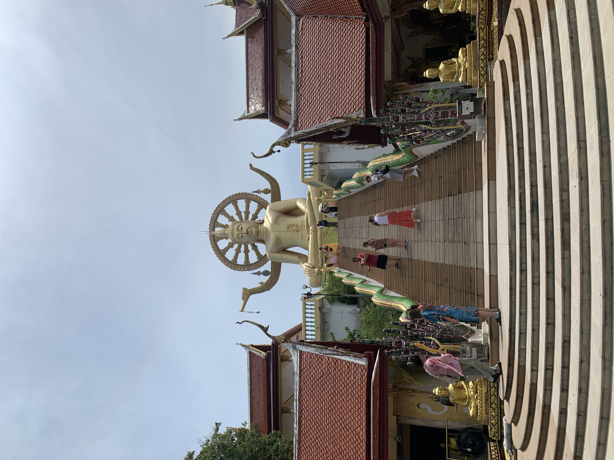 The image depicts the Big Buddha Temple (Wat Phra Yai) in Ko Samui District, Thailand. A colossal golden Buddha statue sits prominently at the top of a wide, tiered staircase. The statue is adorned with a golden halo-like wheel behind its head and has decorative elements extending from its arms.
Numerous people are visible ascending the stairs. Some are walking up, while others are standing or posing for photographs. The stairs are flanked by ornate structures with red tiled roofs and intricate decorative elements, including serpent-like statues with green and gold coloring. Along the sides of the lower stairs, golden Buddha statues are placed on pedestals.
The sky above is overcast with a pale blue hue, suggesting a bright but potentially humid day. Lush green foliage is visible on the sides of the temple structures and behind the main Buddha statue. The overall scene conveys a sense of grandeur and spiritual significance.