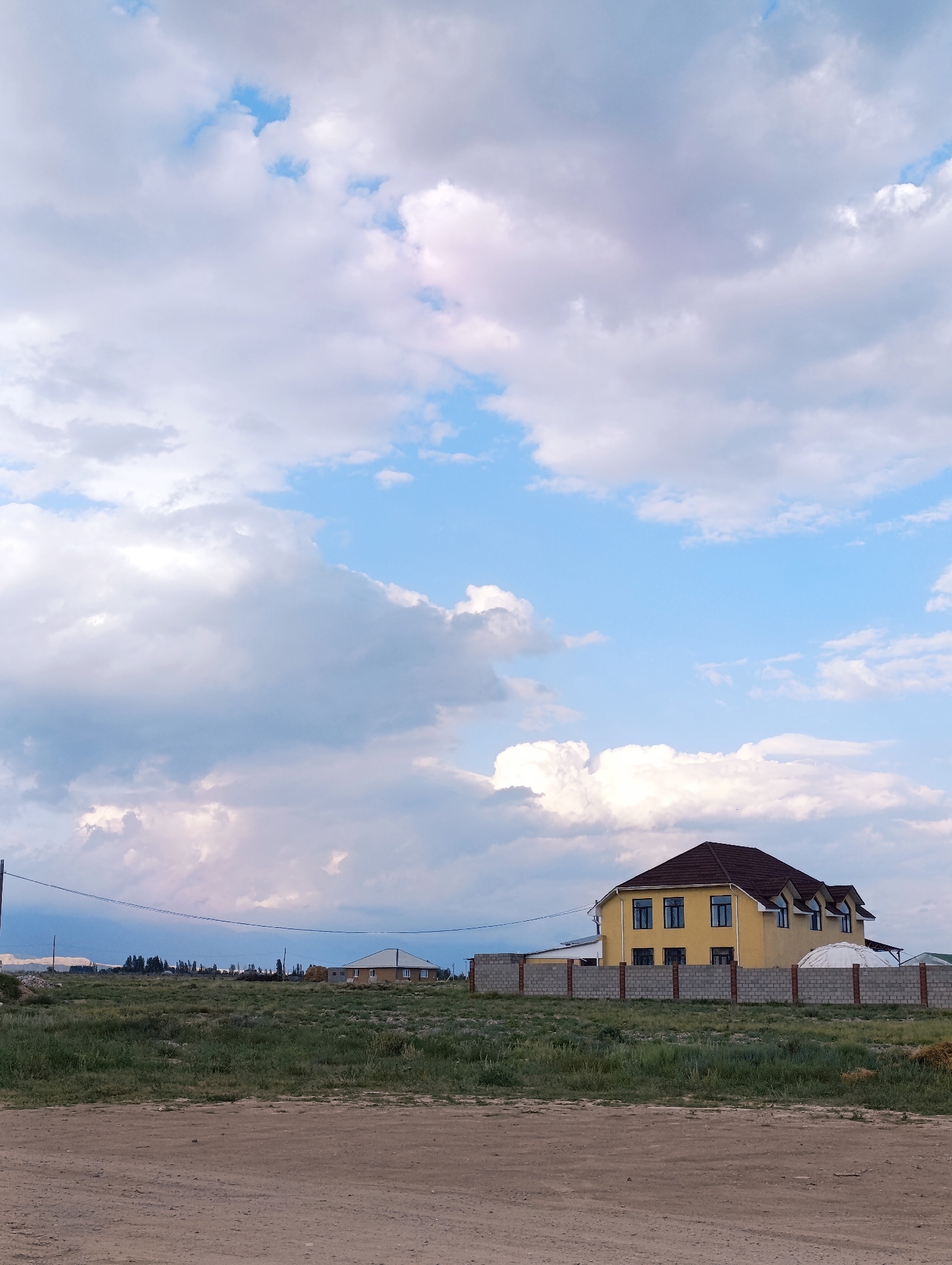 The image depicts a large, two-story yellow house with a dark-tiled roof and dormer windows. The house is surrounded by a low, grey brick and concrete fence. In the foreground, there is a dirt road and an area of dry grass and sparse vegetation. The background shows a flat, open landscape with other buildings and trees visible in the distance, under a sky filled with dramatic white and gray clouds. This scene is located in Toru-Aigyr, Kyrgyzstan.