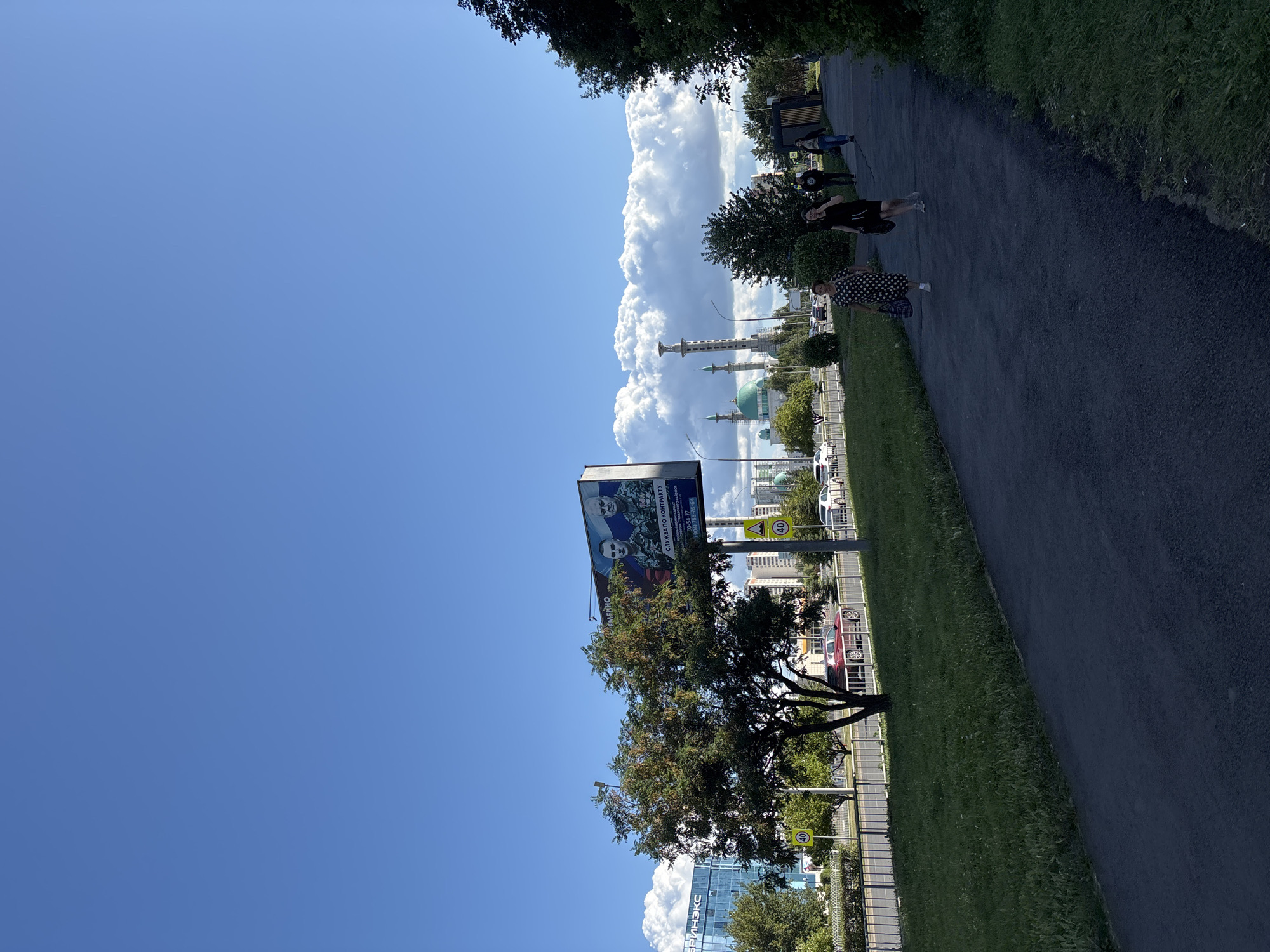 In Naberezhnye Chelny, Russia, on a clear, sunny day with a bright blue sky and scattered white clouds, a paved pedestrian walkway runs alongside a grassy median and a road. Several people are walking on the path. On the road, cars are visible, including a red sedan and a white hatchback. Across the road, a billboard features an advertisement with two military men and Russian flags. Beyond the billboard, a modern blue glass building with the word "БРИНКЭКС" (BRINKEX) is visible. Further in the distance, a mosque with green domes and tall minarets stands against the sky. The environment suggests an urban setting with modern architecture and infrastructure.