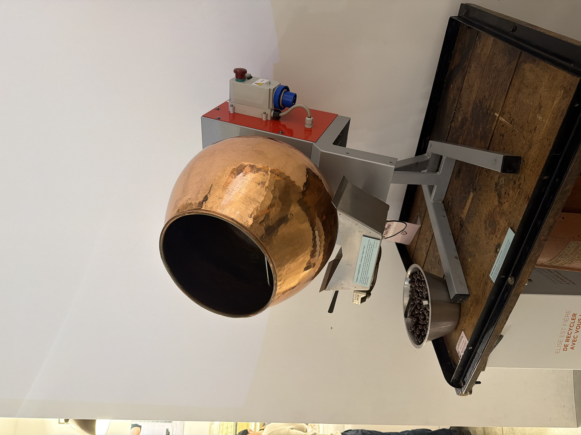 The image shows an exhibition piece in Illkirch-Graffenstaden, France. A copper-colored spherical vessel sits open, angled downwards and to the left. The vessel is attached to a gray metal machine with a red top and a blue electrical connector. A gray metal support arm extends from the machine to a wooden platform with a black metal frame. On the platform, there's a metal bowl filled with brown, oval-shaped objects, possibly coffee beans or chocolate. The background is a plain white wall. Part of a sign with French text, "Elise est fière de recycler avec vous!", is visible on the lower right.