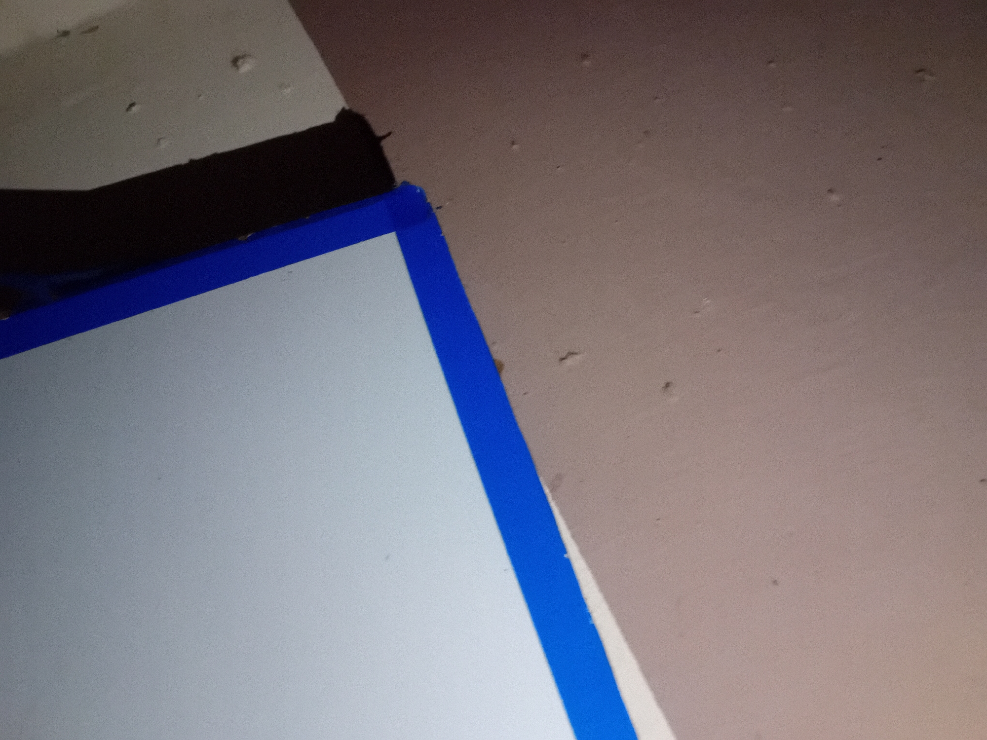 The image shows a close-up view of a corner where a light gray surface, possibly a board or a wall, meets a darker surface, possibly a wall or a floor. A bright blue tape runs along the edge where these two surfaces meet, creating a sharp line. The light gray surface is clean and smooth, while the darker surface has a textured appearance with small bumps or imperfections. The lighting suggests an indoor environment, with a shadow cast by an unseen object or the edge of the gray surface onto the darker surface. The composition is abstract, focusing on the geometric lines and contrasting textures of the materials. The location is an unknown city, and the scene is devoid of any people or discernible activity.