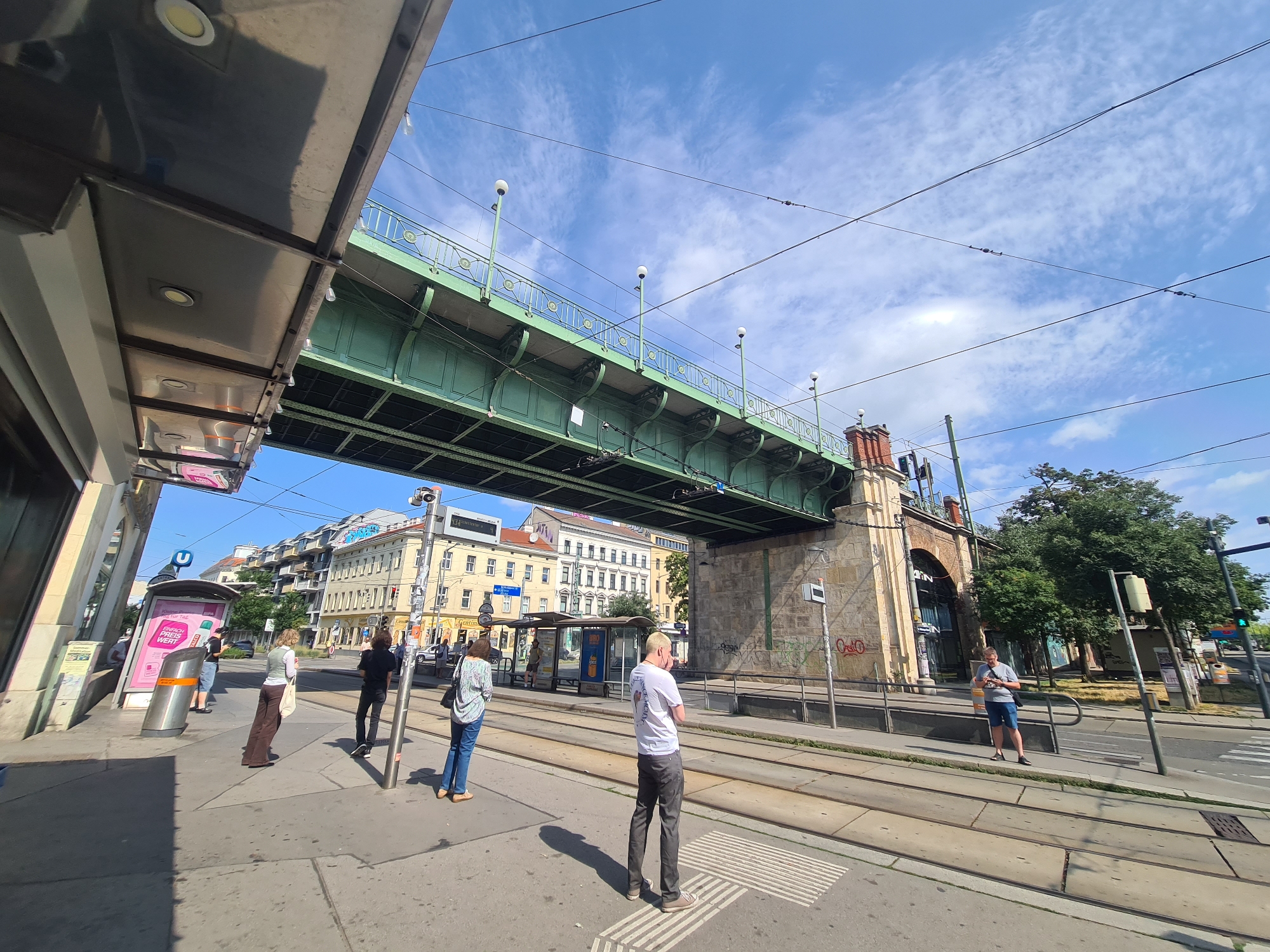 The image depicts a street scene in Vienna, Austria, characterized by an overhead viaduct supporting a green bridge structure. People are visible at a tram stop, waiting for transportation. Several buildings line the street, with some appearing to be older architectural styles, suggesting a historical urban environment. Power lines crisscross the sky, indicating an active transportation system, likely trams or electric buses. The weather appears to be fair with a blue sky and scattered clouds. The scene is captured from a low-angle perspective, emphasizing the scale of the viaduct and the urban infrastructure.