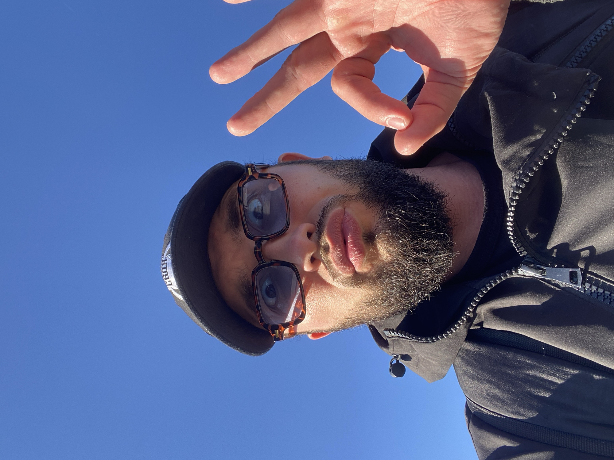 A low-angle, close-up, selfie-style photo taken outdoors against a bright blue sky. The subject, a person with a beard and wearing a black baseball cap and a black jacket, is looking at the camera and making an "okay" hand gesture with their right hand. The person is also wearing thick-rimmed glasses. The background is a clear blue sky, suggesting an outdoor location in the city of Koskolovo, Russia.