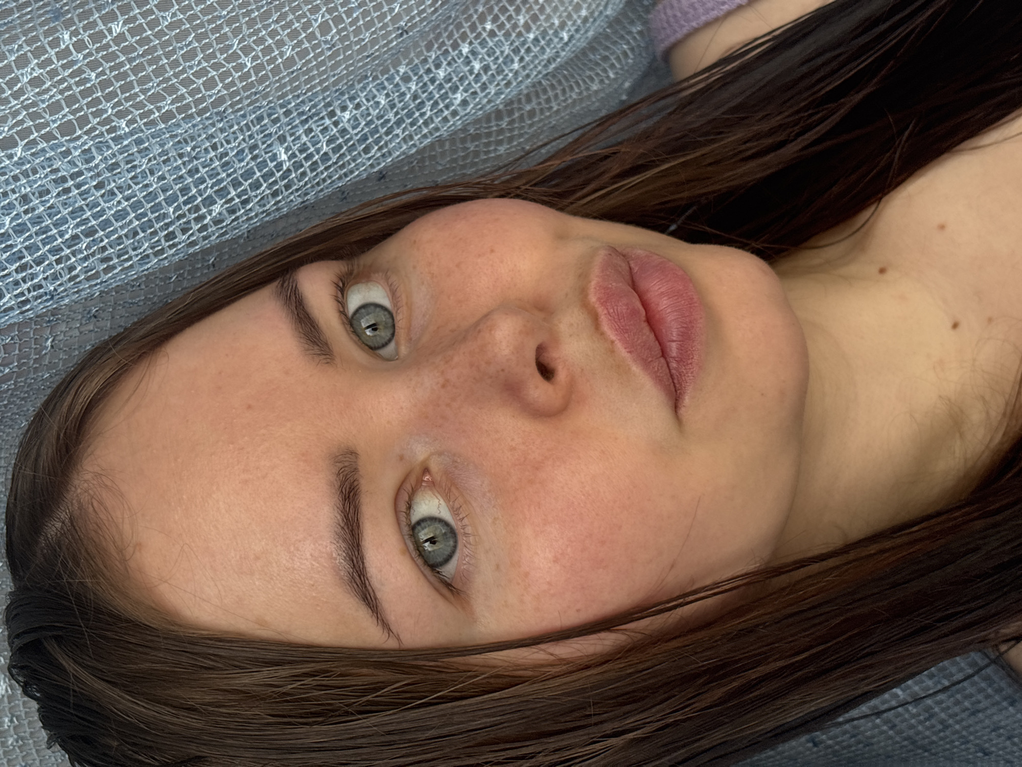A close-up, slightly angled shot depicts a young woman with light skin, freckles, and light blue-green eyes. She has dark brown, wet hair, and her lips are a natural pink color. She is lying down, and a textured blue fabric is visible behind her head. The presence and location of the subject cannot be related to the city of Izhevsk, Russia.