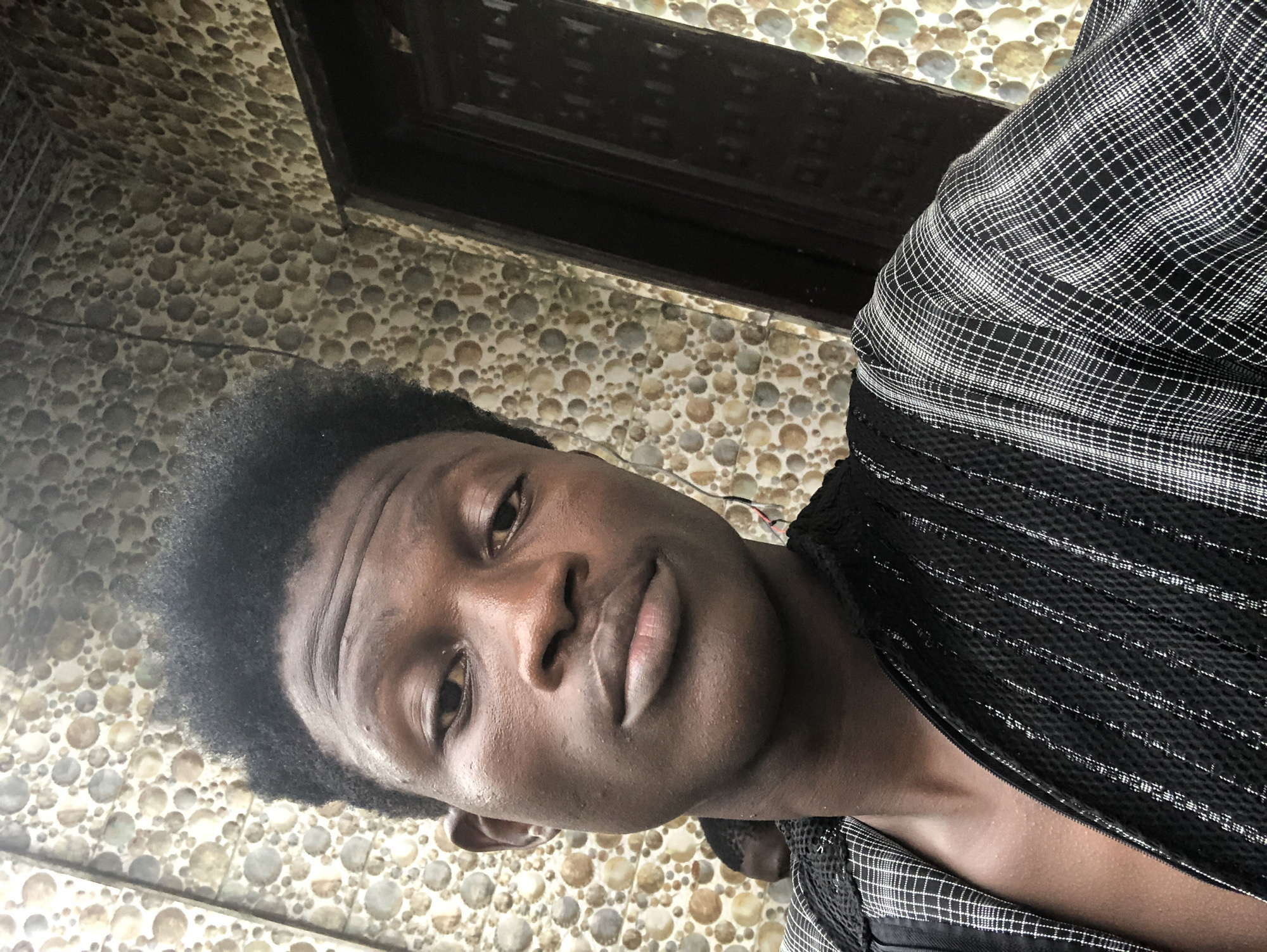 A young Black man is shown in a close-up, selfie-style portrait. He has dark skin, short, dark, textured hair, and is looking directly at the camera with a neutral expression. He is wearing a black and white patterned garment with a zipper. The background consists of a textured wall with a repeating circular pattern, and a dark doorway is visible to the right. The lighting appears to be natural, possibly from an overcast sky. The scene is set in the city of Ajah, Nigeria.