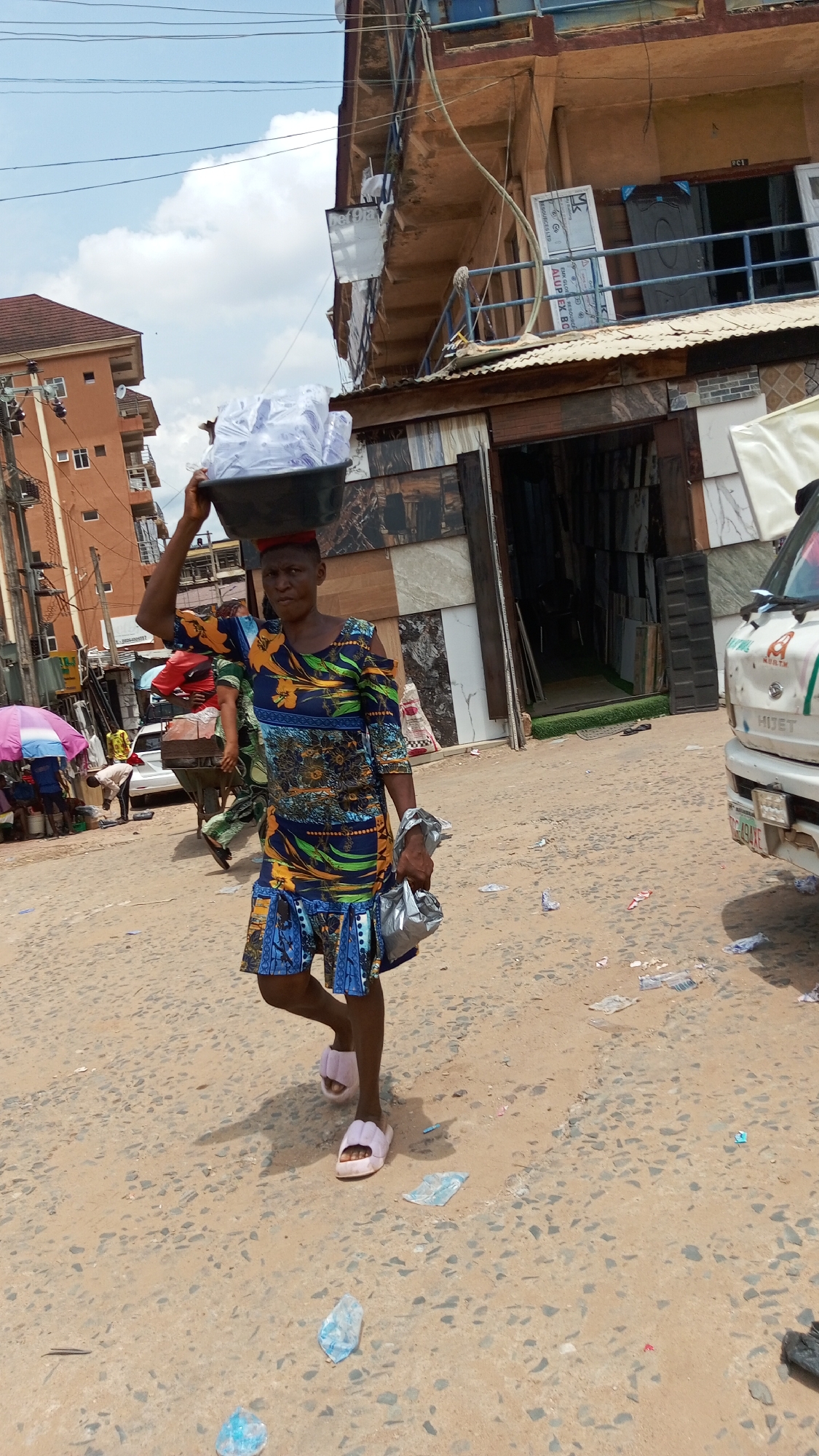 This image depicts a bustling street scene in Ogbunike, Nigeria, under a bright, partly cloudy sky during daylight hours. The main subject is a woman walking towards the viewer, skillfully balancing a large black basin on her head. The basin is filled with numerous clear plastic bags, likely containing sachet water for sale. She wears a vibrant, patterned dress in shades of blue, green, yellow, and orange, featuring cut-out shoulders, and light pink slide slippers. In her left hand, she carries an item wrapped in silver foil.
The street surface is rough and unpaved, littered with scattered empty plastic bags and debris. In the background to the left, another person can be seen pushing a cart under a colorful umbrella. To the right, the front portion of a white van is visible, displaying text such as "KURTM", "HIJET", "ABRTM", and a partial license plate "49AKE".
Buildings line the street, including a multi-story structure with balconies and an adjacent shop. The shop's facade is notably decorated with various samples of wall and floor tiles, suggesting it is a building materials or tile store. Overhead, power lines crisscross the sky. Visible text on signs and banners in the background includes "bet Dia", "RESOURCES LTD", "EMK GLOE RESO", and "ALUP EX BO". The scene captures a typical moment of street commerce and daily life in a West African urban setting. The woman's expression is neutral and focused on her task.