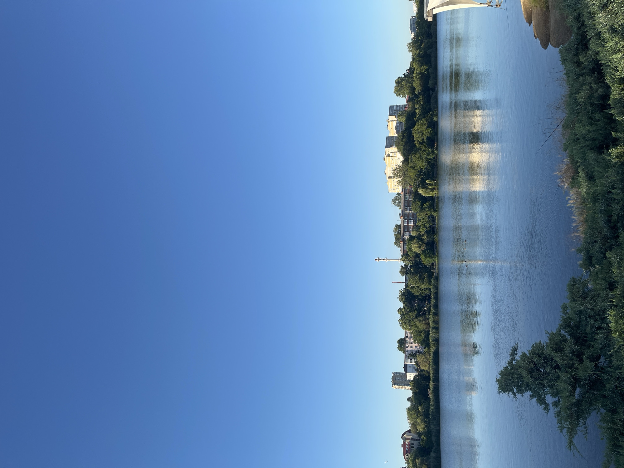The image depicts a serene lake scene with an urban skyline in the background. The sky is a clear, vibrant blue, dominating the upper half of the frame. The lake occupies the mid-ground, its surface reflecting the blue sky and the buildings and trees lining its shores. A few small birds are visible on the water.
In the background, a dense line of green trees separates the lake from a row of buildings. These buildings include a mix of residential structures and what appears to be industrial elements, such as a tall, white chimney with a red top. The architecture suggests an Eastern European urban environment.
The foreground features lush green vegetation and shrubbery, with a prominent evergreen tree on the left. On the right side of the foreground, a white, sail-like structure is partially visible, hinting at a park or recreational area. The overall impression is one of a peaceful coexistence between nature and urban development.
The location is identified as the city of Krasnodar, Russia. The scene likely captures a park or waterfront area within the city, offering a scenic view of the lake and its surroundings. The time of day appears to be morning or late afternoon, given the soft light and the clear sky.
