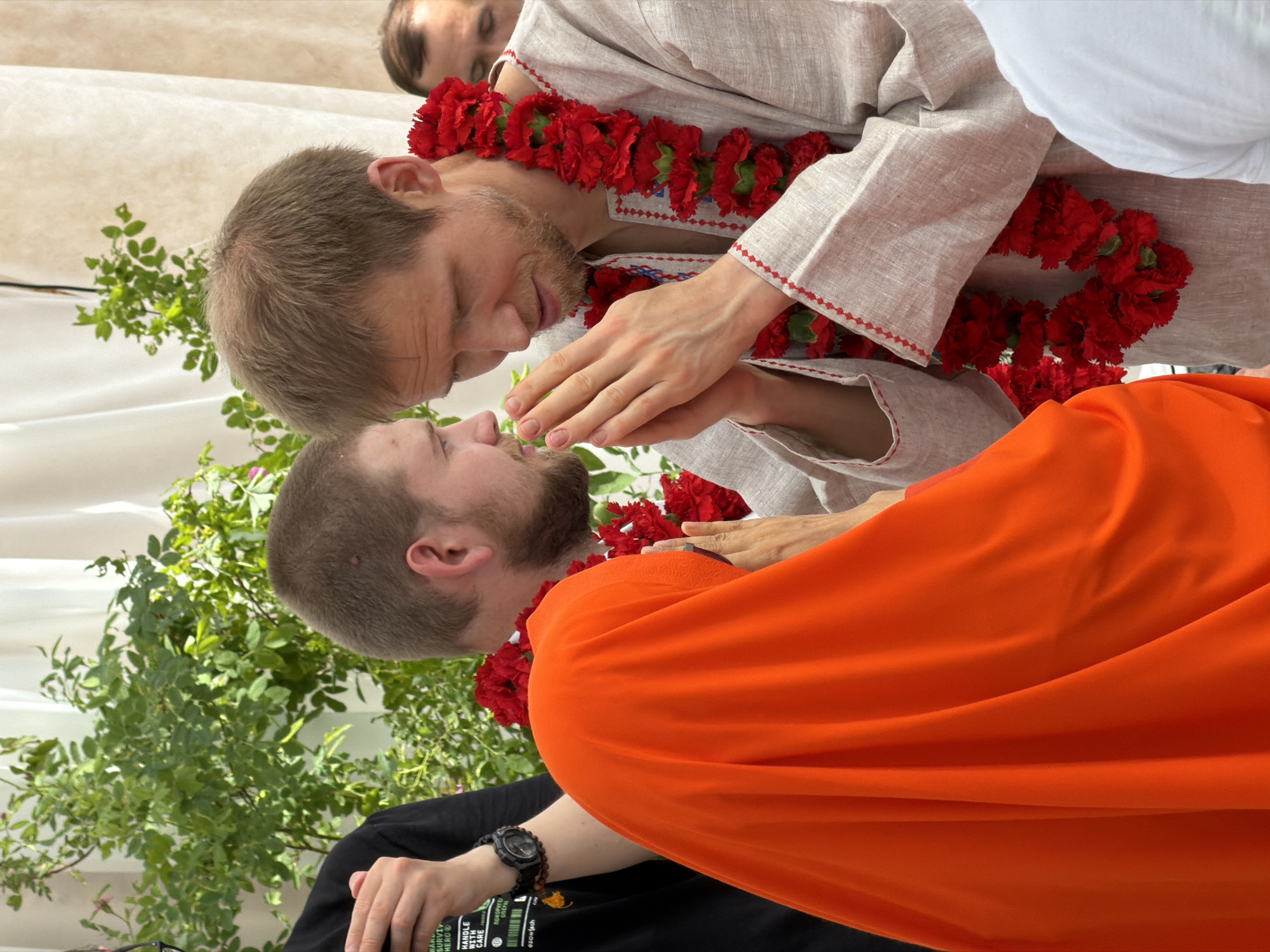 In Zhirnosovo, Russia, three individuals are captured in a moment of respectful interaction. The foreground is dominated by the vibrant orange fabric of a head covering worn by one person, partially obscuring their face and torso. To their right, a man with short, light brown hair and a beard clasps his hands together in a gesture of greeting or prayer. He is adorned with a thick garland of red flowers around his neck. He wears a light-colored, collarless shirt with embroidered details on the neckline and cuffs. To the left of the man with the red garland, another person with short brown hair and a beard is partially visible, also appearing to be engaged in the interaction. In the background, a man with darker hair looks on. Lush green foliage and sheer white drapes form the backdrop, suggesting an outdoor or semi-outdoor setting. The scene appears to be a cultural or religious gathering.