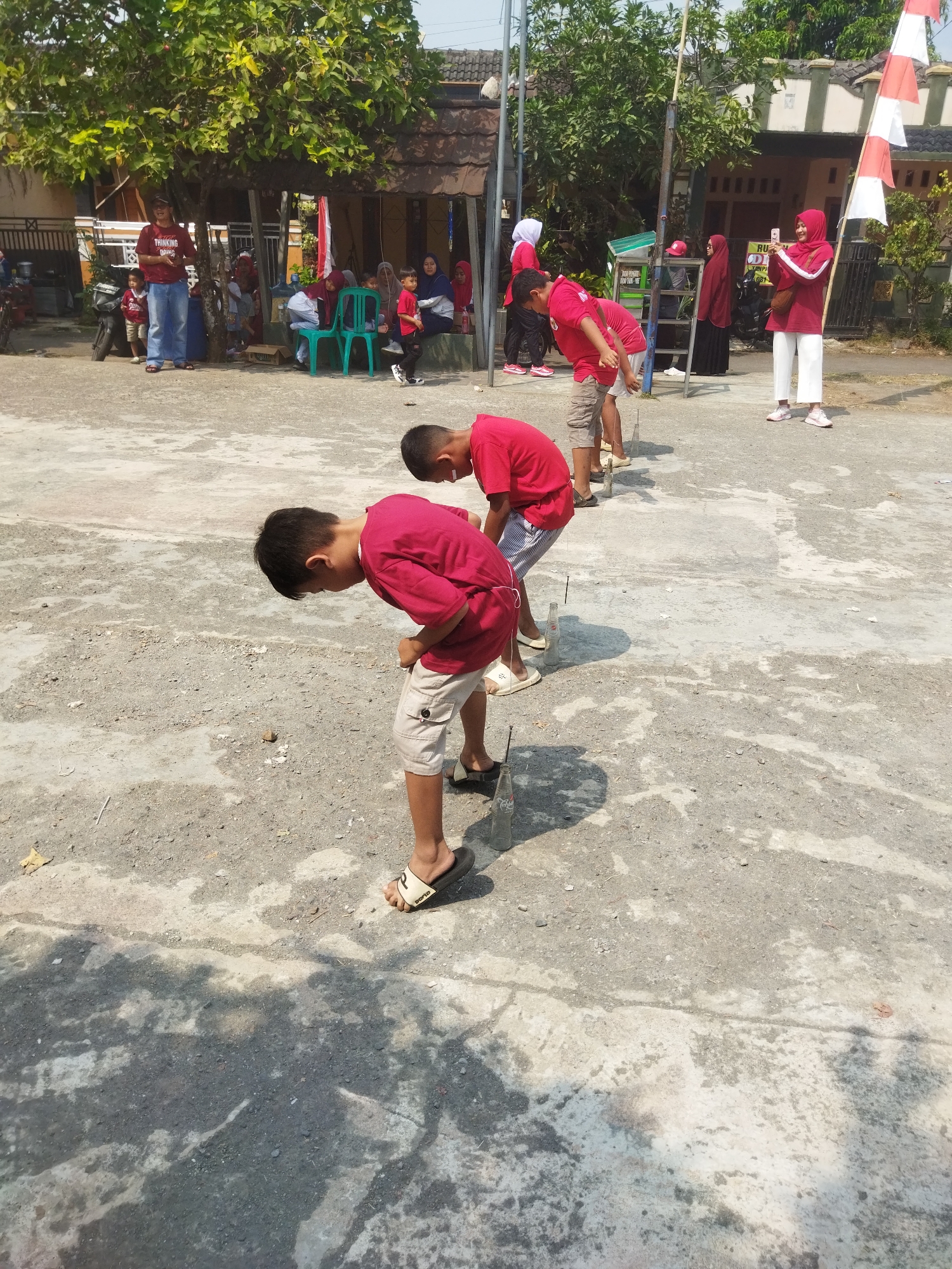 In Banjar, Indonesia, on a sunny day, a group of children are participating in a game that appears to involve placing objects into bottles. The game is taking place outdoors on a dirt and concrete surface, with trees and a building in the background. Several children, mostly boys, are wearing red shirts and are bent over, focusing on the task. Some are holding small items, possibly stones or other game pieces, and aiming them at narrow-necked bottles placed on the ground. Spectators, including adults and other children, are visible in the background, some watching the game and others engaged in conversation or other activities. The environment suggests a casual, community event, possibly a school or local gathering.