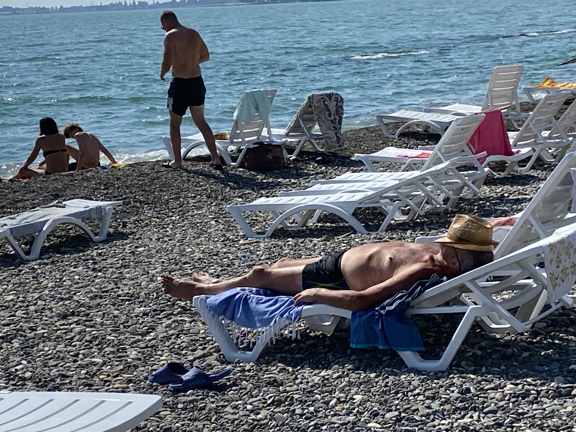 The image depicts a pebbled beach in Chabkwara, Abkhazia, with the sea in the background. Several white plastic lounge chairs are scattered across the beach. In the foreground, a man wearing black swim trunks and a straw hat is lying on a lounge chair, seemingly asleep or resting. A blue towel is draped over his legs. Next to the chair are a pair of blue flip-flops. Further back, near the water's edge, a man in black swim trunks stands, facing away from the camera. Two individuals, a woman in a bikini and a child, are seated on the beach near the water. Several other lounge chairs are visible, some with towels draped over them. The sea is calm and appears to be under a clear sky.