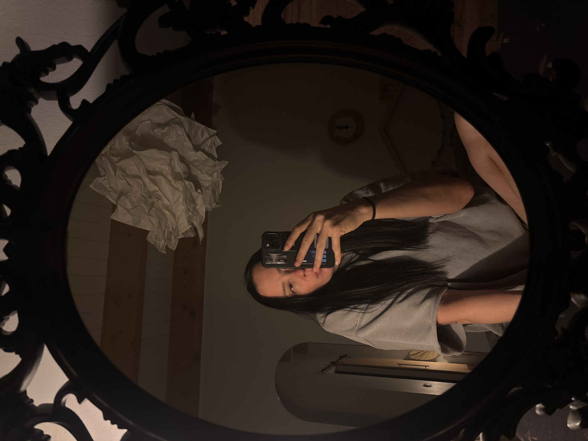 An indoor, low-light selfie captures a young woman reflected in an ornate, dark-framed oval mirror. The woman, with long dark hair, is seen from the chest up, wearing a grey long-sleeved top and a black elastic band on her wrist. She holds a dark-colored smartphone horizontally, pointing its camera towards herself to take the photograph. Her eyes are visible on the phone's screen, looking directly at the camera, while her actual face is partially obscured by the phone.
The reflection in the mirror reveals details of the surrounding room, which appears to be a personal space, likely in Chaikovsky, Russia. In the upper left portion of the reflection, a sizable pile of crumpled white paper or tissues rests on a dark surface. Behind this, light-colored vertical wooden panels are visible. Further in the background, a small, dark, circular object, possibly a clock or decorative item, is reflected high on a wall. The overall dim lighting suggests it is evening or nighttime. Faint text, "NM," is visible on the back of the phone case.