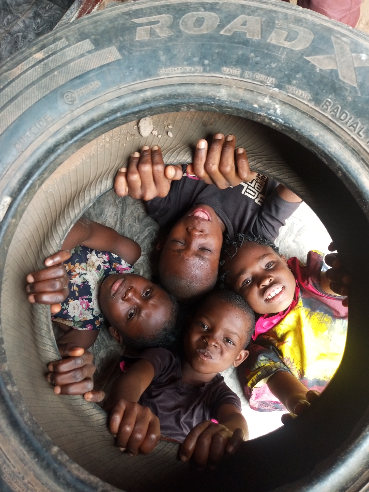 Four children, appearing to be between the ages of 4 and 10 years old, are huddled together inside a large, worn tire. The tire is situated outdoors, on what appears to be a dirt or gravel surface, consistent with an informal setting in a less developed area of Gombe, Nigeria. The children are looking upwards and towards the camera, with various expressions ranging from smiling to curious. Their clothing is casual and colorful. The tire itself shows signs of heavy use and is partially filled with some dry debris. The lighting suggests it is daytime. The scene conveys a sense of playfulness and camaraderie among the children despite the humble surroundings.