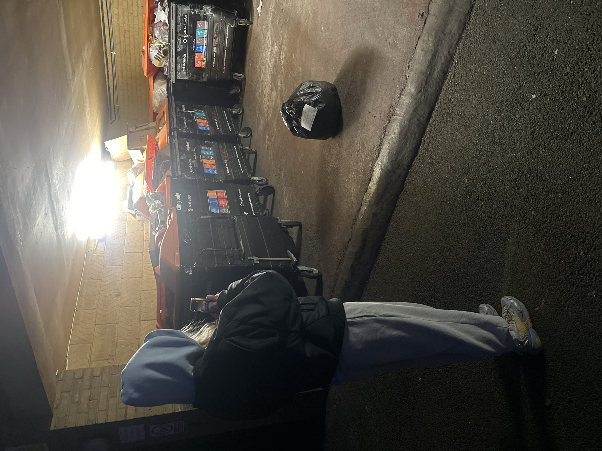 The image shows a person in grey sweatpants, a black jacket, and a blue hoodie, holding a smartphone and facing several rows of large, black, wheeled recycling bins. A black trash bag sits on the ground between the rows of bins. The environment appears to be an alleyway or loading area, possibly at night, with a light source causing glare in the upper left corner. Markings on the recycling bins indicate "Hackney" and "Recycle for London", suggesting the location is in the Hackney area of London, United Kingdom. The bins are labeled with recycling instructions, including "cling only" and "No bl dkbags." The scene is lit unevenly, with some areas appearing dark.