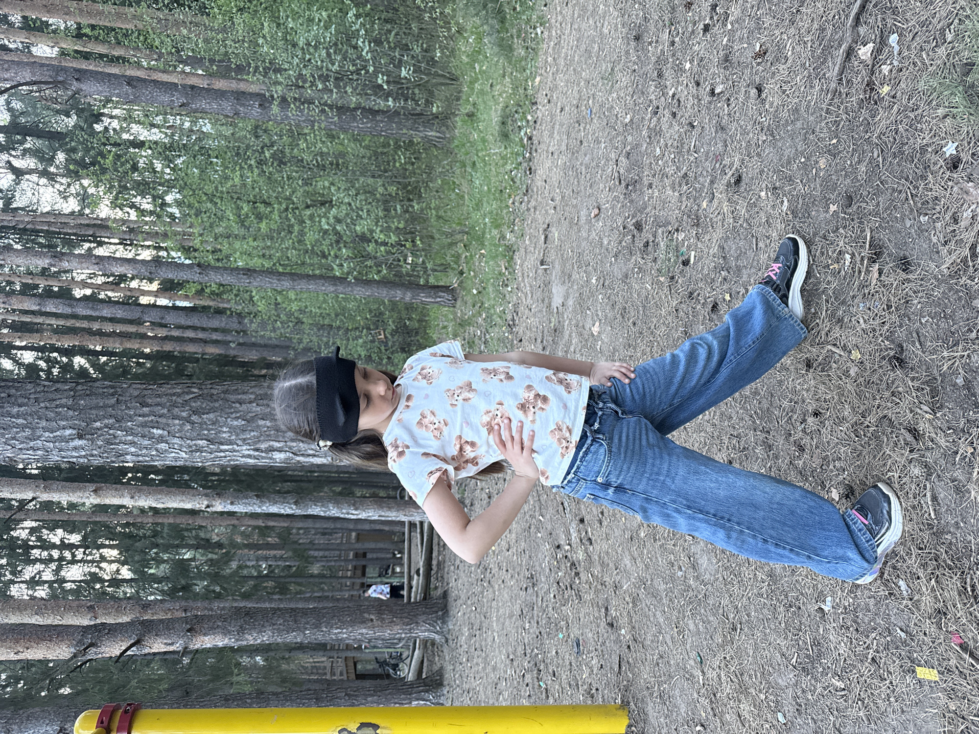 A young girl with a black blindfold poses outside in what appears to be a park setting in Votkinsk, Russia, as suggested by the vegetation, infrastructure, and typical park scenery. She wears a white T-shirt with a light brown teddy bear pattern, blue jeans, and black and white sneakers. The ground around her is a mix of dirt, grass, and scattered debris. Trees are visible in the background, creating a forest-like atmosphere. A yellow metal bar is in the lower left corner. The interaction is between the girl and the camera, as she appears to be posing for a photograph.