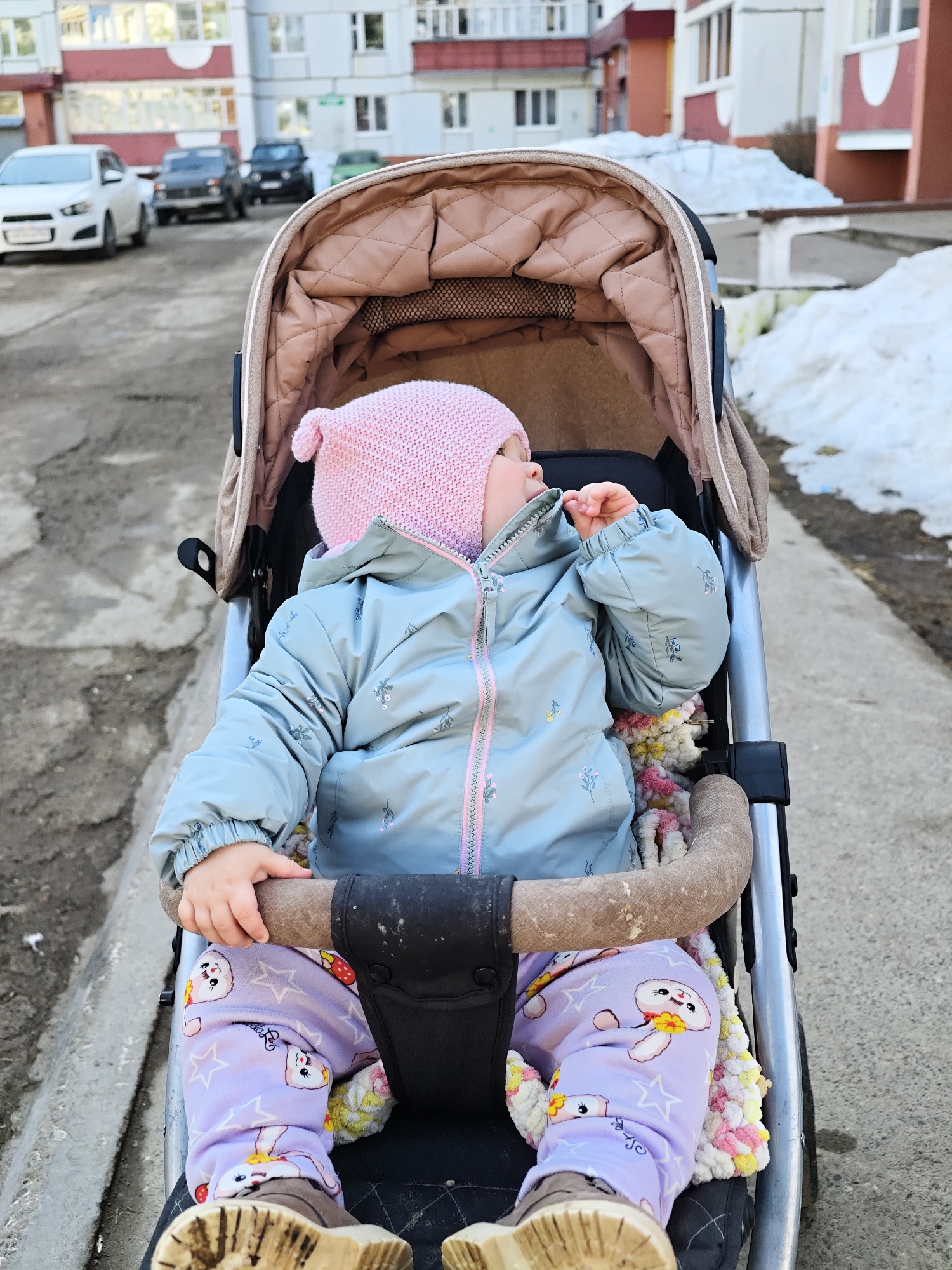 A small child in a stroller is centered in the frame, wearing a pink knit hat, a light blue quilted jacket, and patterned purple pants. The child has their hand on the stroller's safety bar and appears to be looking upwards. The stroller is on a concrete sidewalk. Behind the stroller, there are cars parked on a street and buildings, typical of urban architecture. To the right of the stroller, partially melted snow sits on the side of the sidewalk. The image appears to be taken in Chaikovsky, Russia.