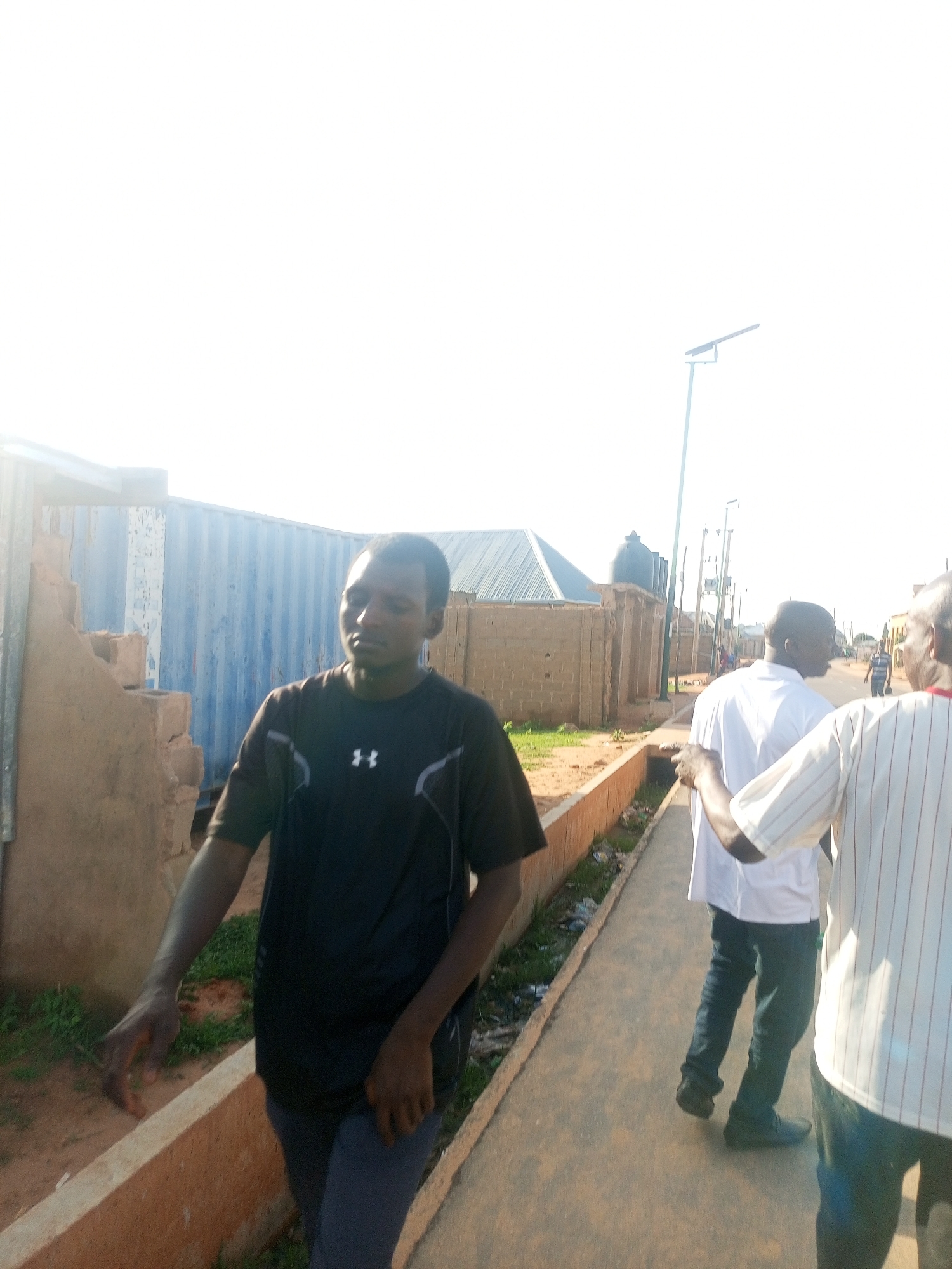 This image shows an outdoor scene in Tempure, Nigeria. A man in a dark t-shirt and dark pants is in the foreground, with his left arm extended and his hand resting on a low wall. Behind him, the wall transitions into a blue corrugated metal structure. To the right, two other men are walking away from the camera. One is wearing a white shirt and dark pants, and the other is wearing a striped shirt. They are on a paved walkway. In the background, there are buildings made of brick and corrugated metal, along with streetlights. The sky is bright and appears to be daytime.