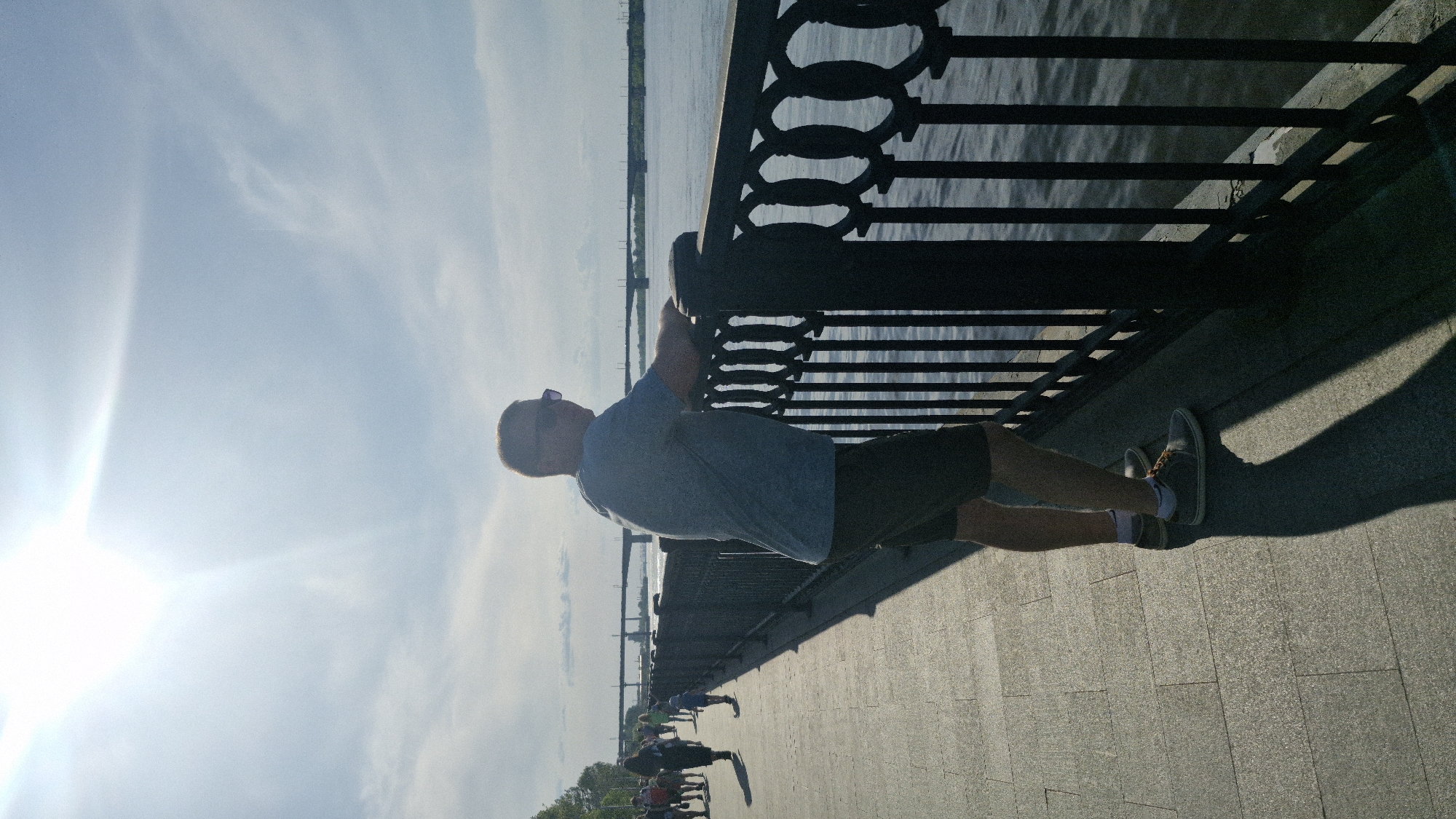 A man in a blue t-shirt, dark shorts, and sneakers with white socks is seen holding onto a dark metal fence, possibly doing a stretching exercise. The fence runs along a paved walkway next to a body of water. In the distance, other people are walking along the walkway. The sky is bright with the sun visible and some light clouds. The photo was taken at the Perm embankment along the Kama River in Perm, Russia.