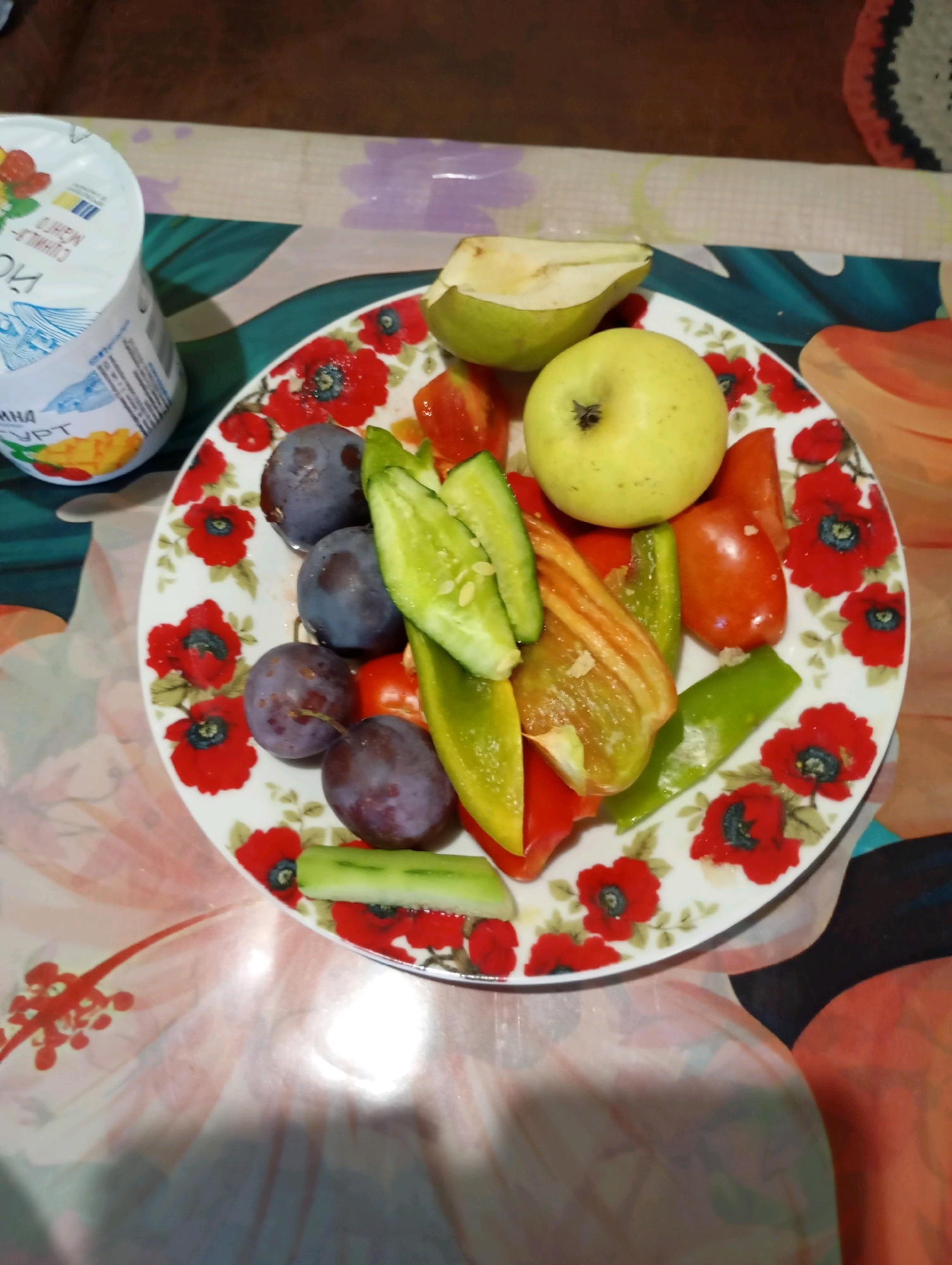 A close-up overhead shot shows a plate of cut fruits and vegetables on a floral patterned tablecloth. The plate is white with a red poppy design. On the plate, there are several whole purple plums, slices of cucumber, wedges of bell peppers (red and green), small red tomatoes, a sliced pear, and a whole green apple. To the left of the plate, a partially visible container of yogurt with fruit imagery is present. The overall scene suggests a meal or snack preparation. The location is identified as Horishni Plavni, Ukraine.