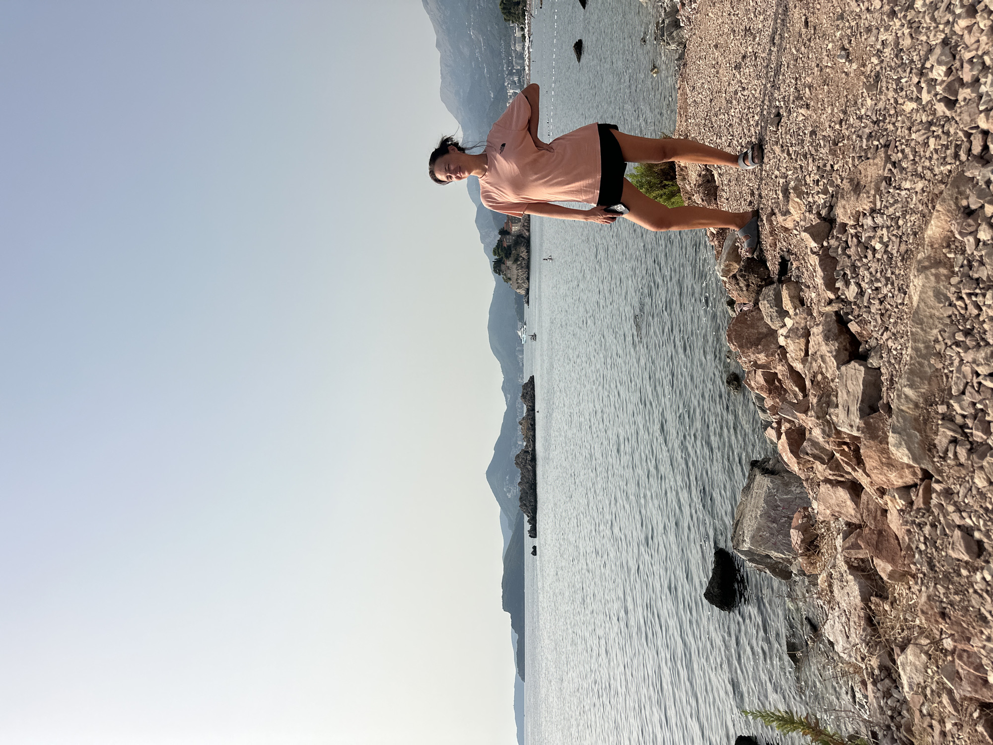 In Sveti Stefan, Montenegro, a woman stands on a rocky shore with a view of the Adriatic Sea. The woman is wearing a peach-colored t-shirt and black shorts, with her hand on her hip. She is looking to her left, with a slight smile on her face. In the background, the sea stretches out towards distant islands and mountains, with buildings visible along the coastline. The sky is clear and light blue, with a hazy quality. The foreground is composed of various sizes of rocks and pebbles.