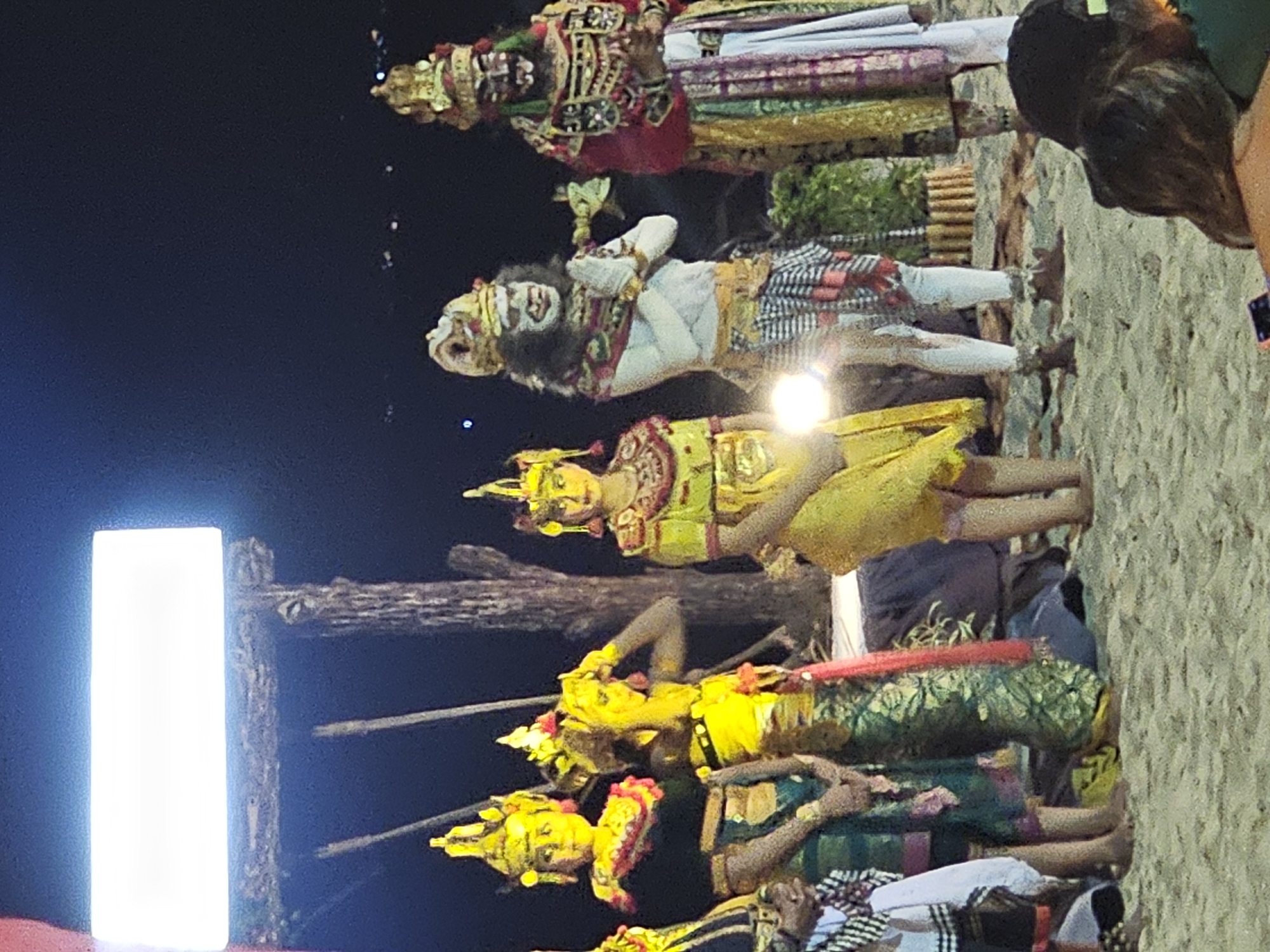 The image captures a performance of a Balinese dance, likely a Ramayana ballet, in Denpasar, Indonesia. The dancers are adorned in vibrant traditional costumes, including elaborate headdresses and face paint, with details in gold, yellow, green, red, and white fabrics. They are positioned in a line, suggesting a segment of a choreographed routine. The stage appears to be set outdoors, near a light source that casts a stark illumination, and possibly against a simple rustic wooden backdrop. A small audience, including at least one person with dark hair, can be seen in the periphery.
