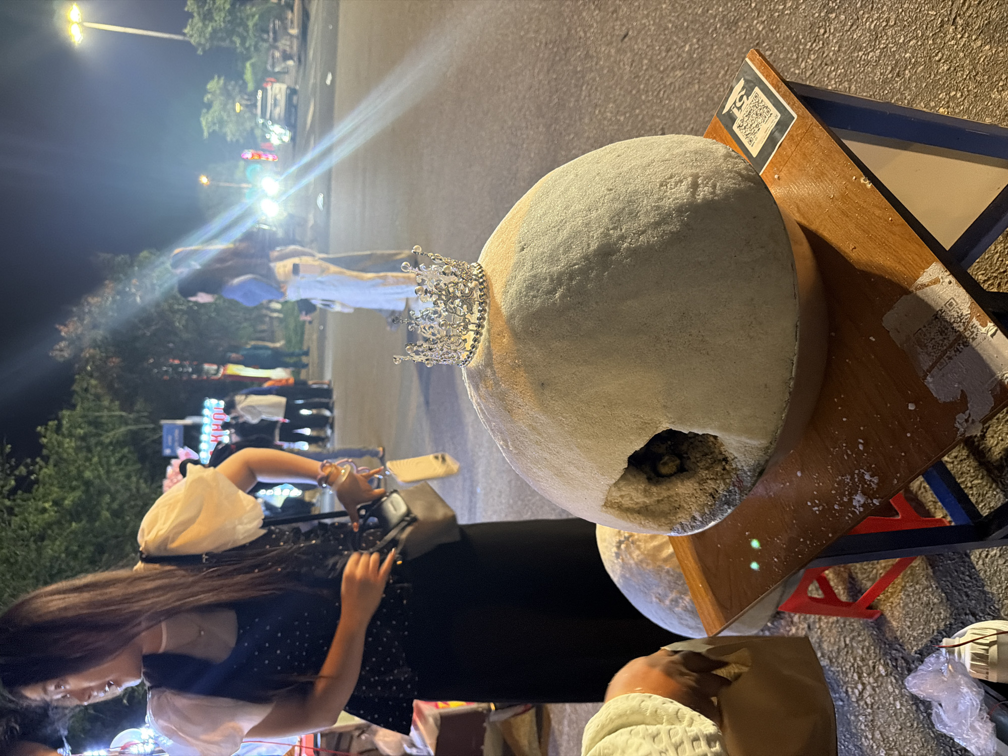 This image captures a vibrant night market scene in Măng Đen, Vietnam. The central focus is a large, rough-textured spherical object, possibly made of clay or concrete, positioned on a wooden stand. A small, elaborate silver crown rests atop this sphere, which also features a small opening on its side revealing a dark interior. A smaller, similar spherical object is partially visible beneath it.
To the left, a woman with long, dark hair is seen from the side, wearing a dark polka-dotted dress over a white puff-sleeved top. She is engaged with items on the stand, possibly interacting with a mobile phone or another device, with her hands poised. Another person, partially visible in the bottom right, appears to be seated or kneeling beside the stand.
The setting is outdoors at night, with the sky dark and various bright artificial light sources, including streetlights and what appear to be vehicle headlights, creating strong glares and light streaks across the frame. In the blurred background, other market-goers, cars, and trees are discernible. The wooden stand supporting the main sphere has two QR codes affixed to it; one is clearly visible, while the other is partially obscured and worn. Miscellaneous items like plastic bags and wires are visible on the ground around the stand. The overall atmosphere suggests a lively commercial street or market environment after dark.