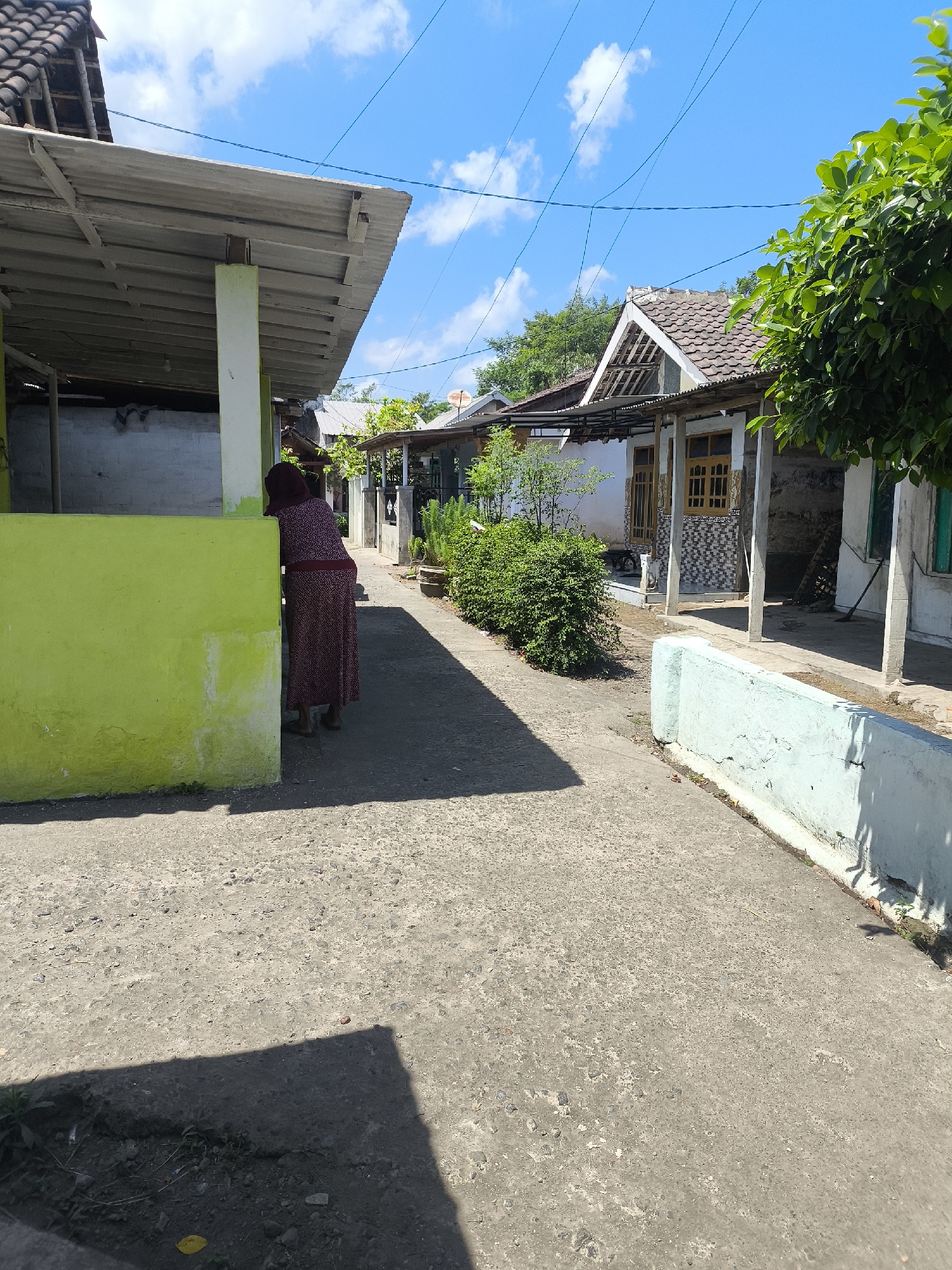 The image depicts a residential alleyway in Grati, Indonesia. The scene is dominated by a concrete pathway leading into the distance, bordered by simple houses with tiled roofs and covered verandas. On the left, a person wearing a maroon patterned dress and headscarf leans against a bright lime green wall, partially obscured from view. Sunlight casts shadows across the pathway, indicating a bright day with a clear blue sky dotted with white clouds. Lush green foliage is visible on the right side of the alley and in the background, suggesting a tropical climate. The houses exhibit a mix of architectural styles, with some featuring white walls and yellow-framed windows, while others appear more weathered. The overall impression is of a quiet, sun-drenched neighborhood.