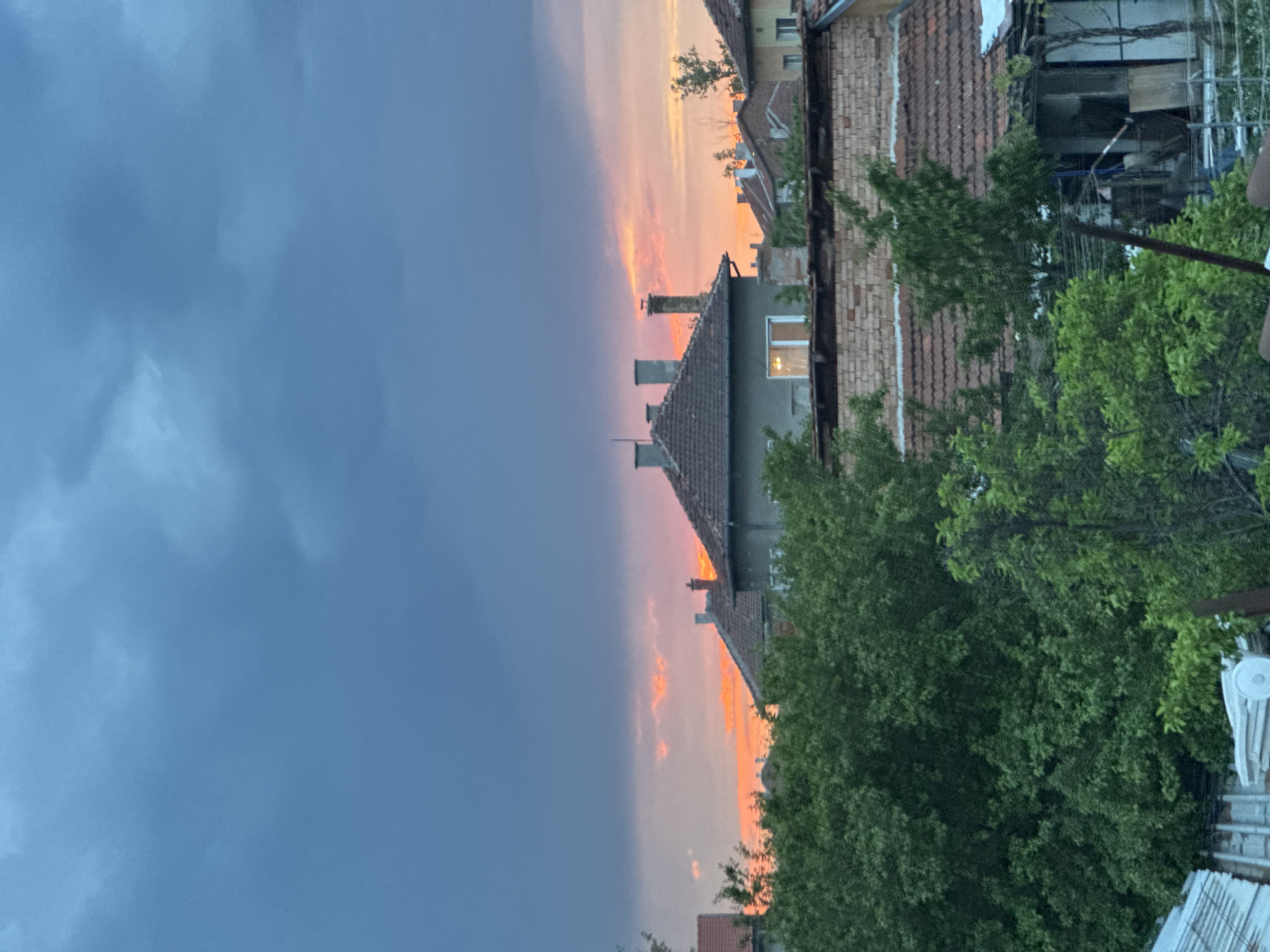 The image shows a view from what appears to be a residential area, possibly in Elin Pelin, Bulgaria. The sky is divided with a dark blue, ominous cloud formation occupying the left side of the frame, indicative of an approaching storm or recent rainfall. On the right, there's a vibrant sunset with hues of orange and pink coloring the sky. The sunset illuminates the rooftops of several buildings, one with multiple chimneys, and highlights the foliage of a green tree. There are a number of buildings that appear to be 1-2 story residential homes, typical of Elin Pelin. The foreground includes green trees and glimpses of fencing and construction materials, indicating an inhabited or in-progress area. There are no visible people or animals.