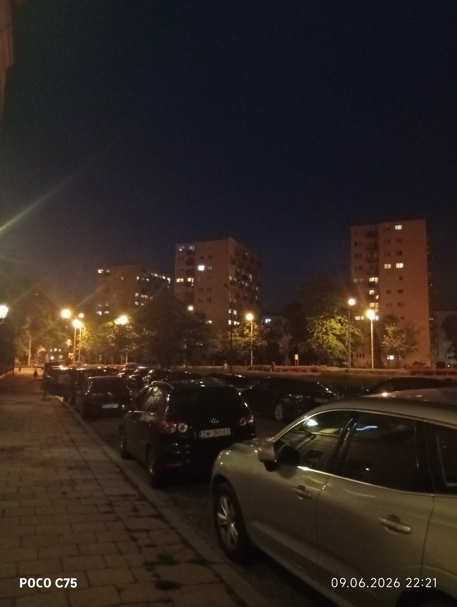 A nighttime street scene in Wrocław, Poland, features a row of parked cars along a brick sidewalk. In the background, several multi-story apartment buildings are illuminated with interior lights, and trees are visible between the buildings and the street. Streetlights cast a warm glow on the scene. The image also includes a timestamp indicating it was taken on 09.06.2026 at 22:21 and a device identifier "POCO C75". No people are clearly visible in the frame.