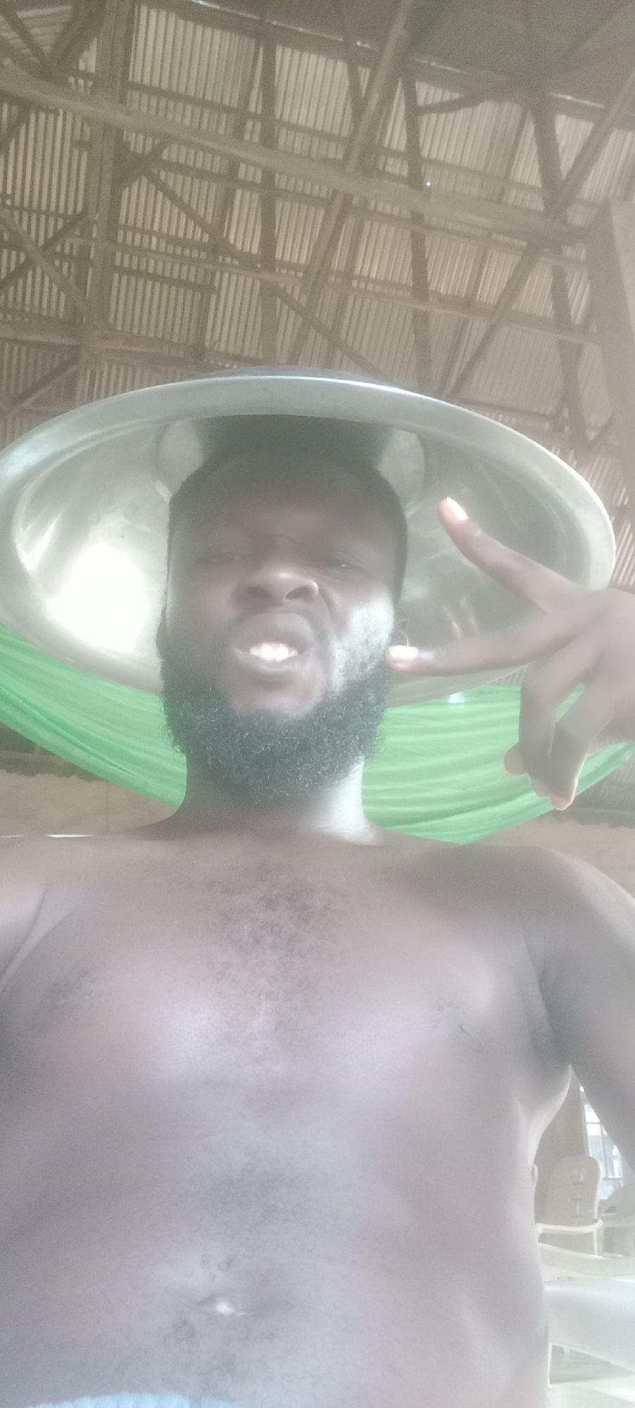 A shirtless, dark-skinned man with a beard and a metal bowl on his head is posing for a selfie. He is making a peace sign with his right hand and grimacing. In the background, there is a green hammock and a corrugated metal roof supported by wooden beams. The image was likely taken in a building in Bende, Nigeria.