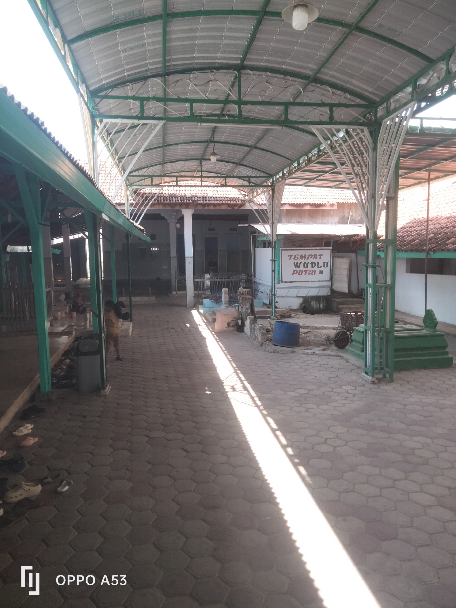 The image depicts an outdoor covered walkway with a hexagonal tiled floor. The walkway has a green metal roof supported by white metal beams, some of which are ornately decorated. Sunlight streams through the gaps in the roof, casting long shadows on the ground.
In the background, a building with tiled roofs and white walls is visible. A sign with red text on a white background reads "TEMPAT WUDLU PUTRI," indicating a women's ablution area. To the right, a green tomb or shrine is present.
Several pairs of shoes are scattered on the tiled floor near the walkway. A trash can is also visible. In the mid-ground, a young person in a yellow shirt and dark shorts stands near the trash can. Further to the left, an adult is seated. In the distance, other individuals are present, as well as what appear to be more graves or resting places. The overall environment suggests a place of worship or historical significance, possibly a cemetery or mosque complex.