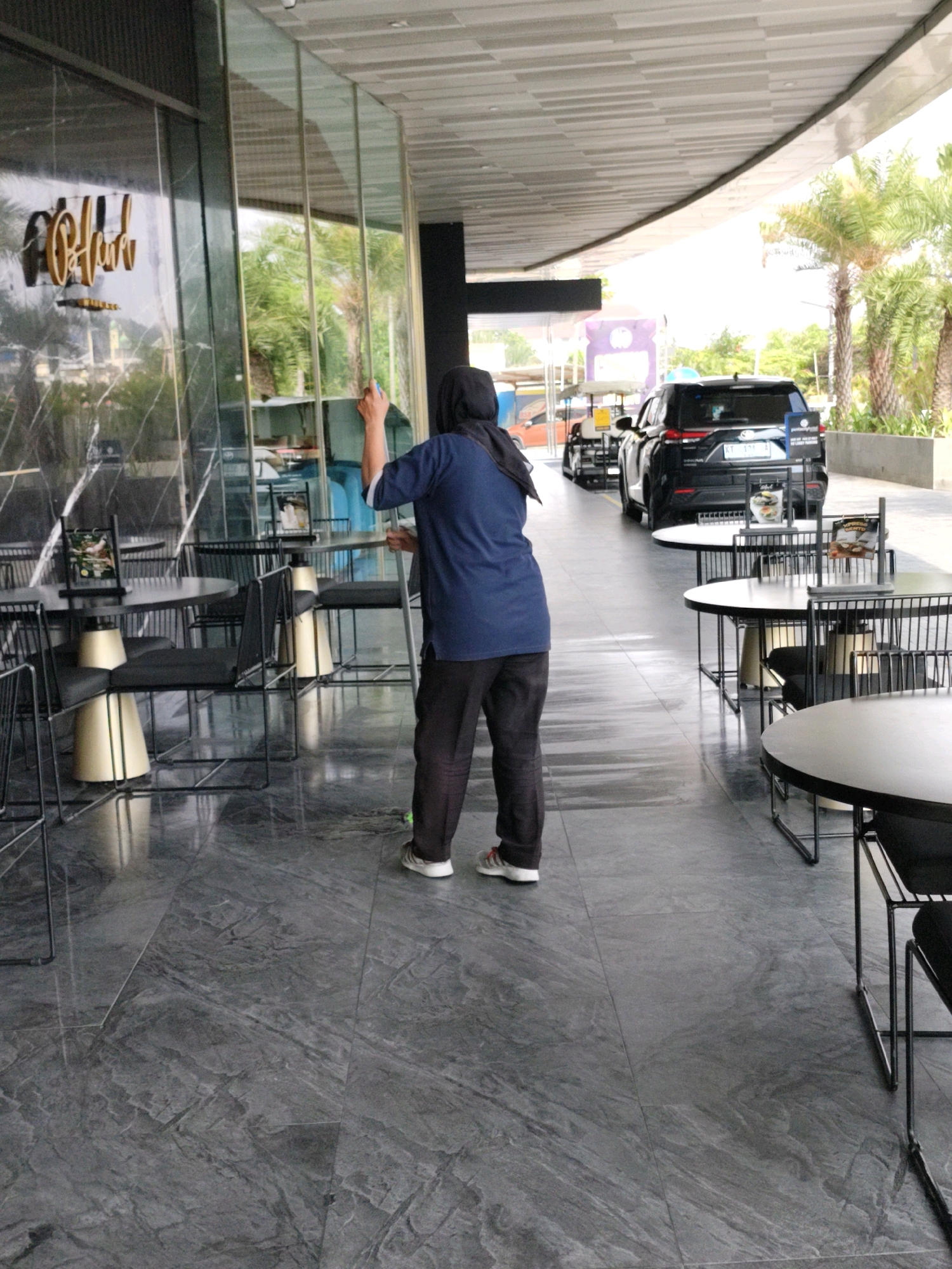 A person wearing a dark blue shirt, dark pants, and a black headscarf is mopping the tiled floor in front of a building with a modern facade. The building has large glass windows, and a sign with golden lettering is visible on one of them. Outside, several outdoor dining tables and chairs are arranged on the walkway. In the background, a black SUV and a few golf carts are parked. Palm trees are visible in the distance. The scene appears to be a public area, possibly a cafe or restaurant entrance, in the city of Balikpapan, Indonesia.