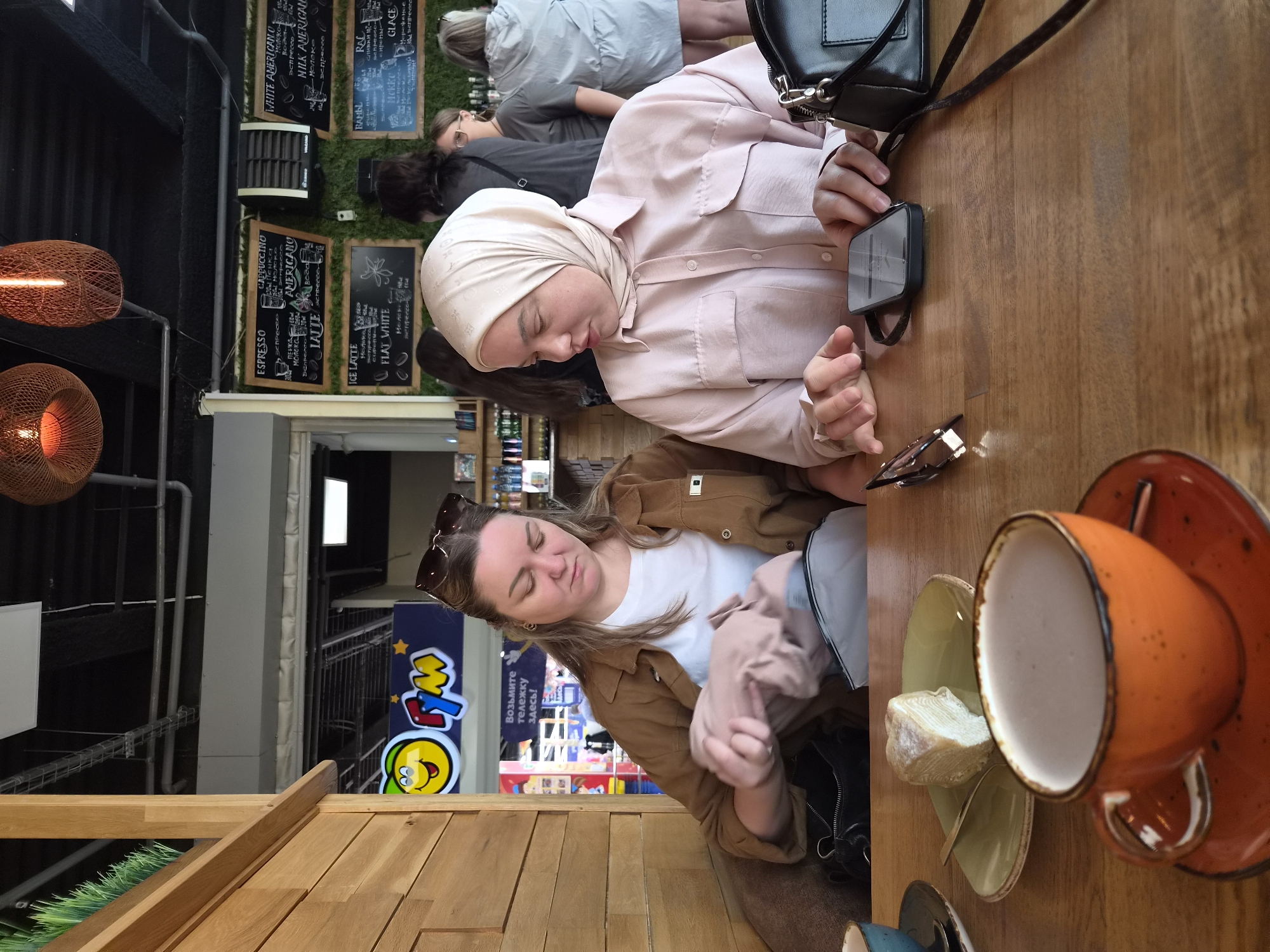 The image depicts two women seated at a wooden table in an indoor cafe. The woman on the left, wearing a brown jacket over a white t-shirt, is rummaging through a light-colored bag. She has dark hair and is looking down at the bag. The woman on the right is wearing a pale pink hijab and a matching button-down shirt. She is looking at a smartphone placed on the table in front of her, with her hands resting on the table.
On the table, between the two women, is a slice of layered cake on a green plate with a fork, and an orange coffee cup filled with a light-colored beverage. To the right of the woman on the right, a black handbag rests on the table.
The background reveals a cafe environment with wooden paneling on the left and a wall with chalkboards displaying a menu on the right. The chalkboards list various coffee drinks and prices. In the distance, other patrons and a counter area are visible. The cafe appears to be part of a larger establishment, possibly a shopping center, suggested by signage in Russian. One sign, featuring a cartoon character, reads "Возьмите тележку здесь!" which translates to "Take a trolley here!". This suggests the location is within a shopping area, and potentially the Tsentralnyi Rynok in Arseniev, Russia, given the style of signage and the mention of trolleys often found in markets. The overall lighting is warm, typical of an indoor dining setting.