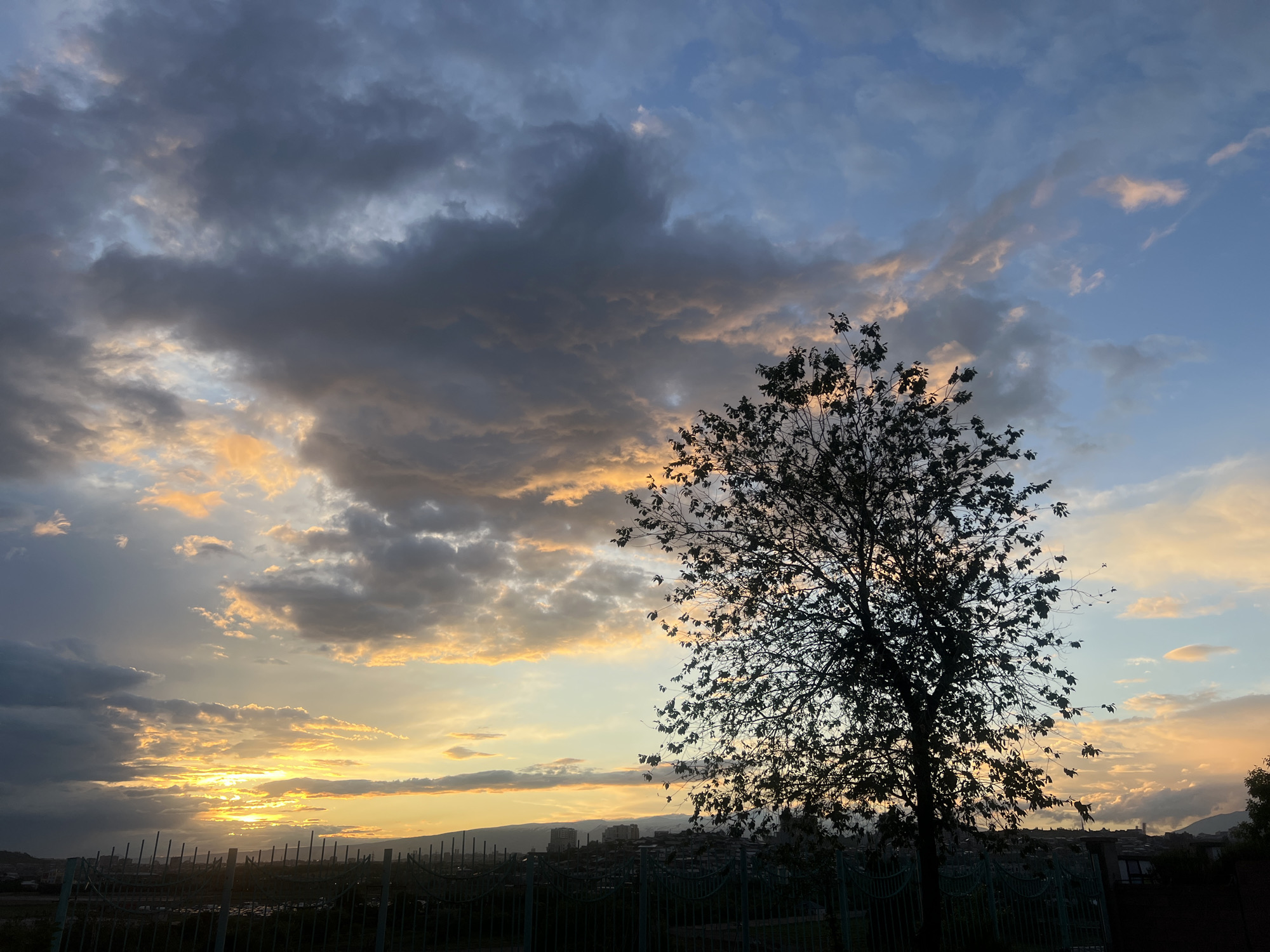 A wide-angle shot captures a sunset scene in Yerevan, Armenia. The sky features a mix of dark, gray clouds and areas of clear, light blue sky with orange-tinged clouds near the horizon. A silhouetted tree stands in the right-center foreground. In the midground, a dark metal fence runs horizontally across the frame. Beyond the fence, a blurred urban landscape is visible, including buildings and distant hills. The sunset is positioned on the left side of the frame, casting orange light across the horizon.
