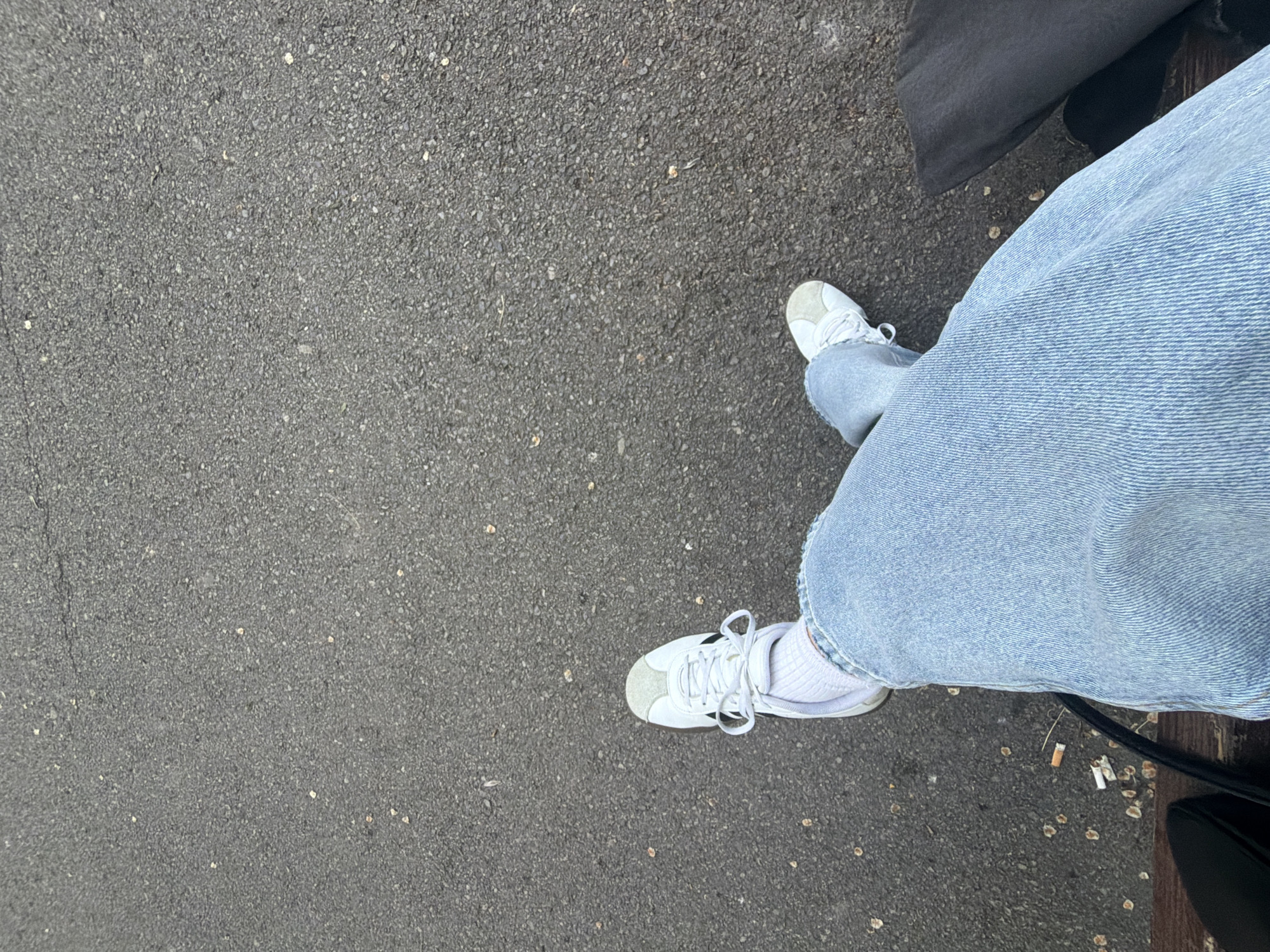 The image shows a view from above, looking down at a pair of legs in light-wash denim jeans and white sneakers. The person appears to be sitting, as a small portion of a black bag and what looks like a bench are visible. The ground is asphalt. There are cigarette butts, and small debris scattered on the asphalt. There is no indication in this image of it being taken in Sofia, Bulgaria. No actions or interactions are visibly depicted.