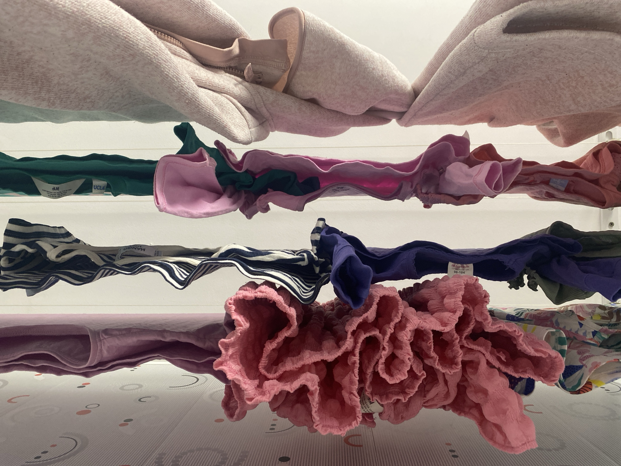The image depicts multiple articles of clothing suspended from a drying rack. The garments vary in color and style, including pink, green, blue, and striped patterns. Some items appear to be children's clothing. Labels from brands like H&M and MANGO are visible on some items. The drying rack is positioned indoors, as evidenced by the presence of a patterned surface underneath. The location of the image is not ascertainable from the provided visual data.