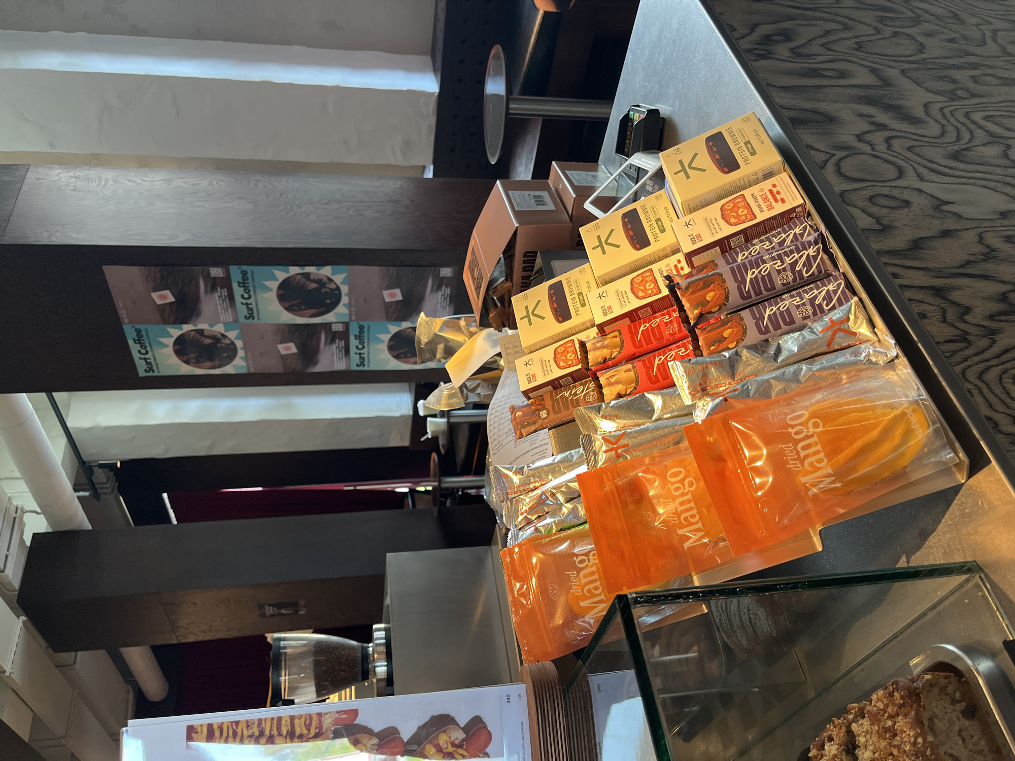 The image depicts a retail counter inside a cafe, likely in Kazan, Russia, featuring a variety of packaged food items. Dominating the foreground are several orange pouches labeled "dried Mango" and various chocolate bars and protein bars in colorful packaging. To the left, a display case contains a slice of cake topped with nuts. In the background, a coffee grinder is visible behind a menu board. The counter itself is made of dark wood with a visible grain pattern. In the far background, dark red curtains and upholstered seating suggest a dining or lounge area. A circular table and chairs are also visible, along with a payment terminal on the counter. The overall environment appears to be a modern cafe setting.