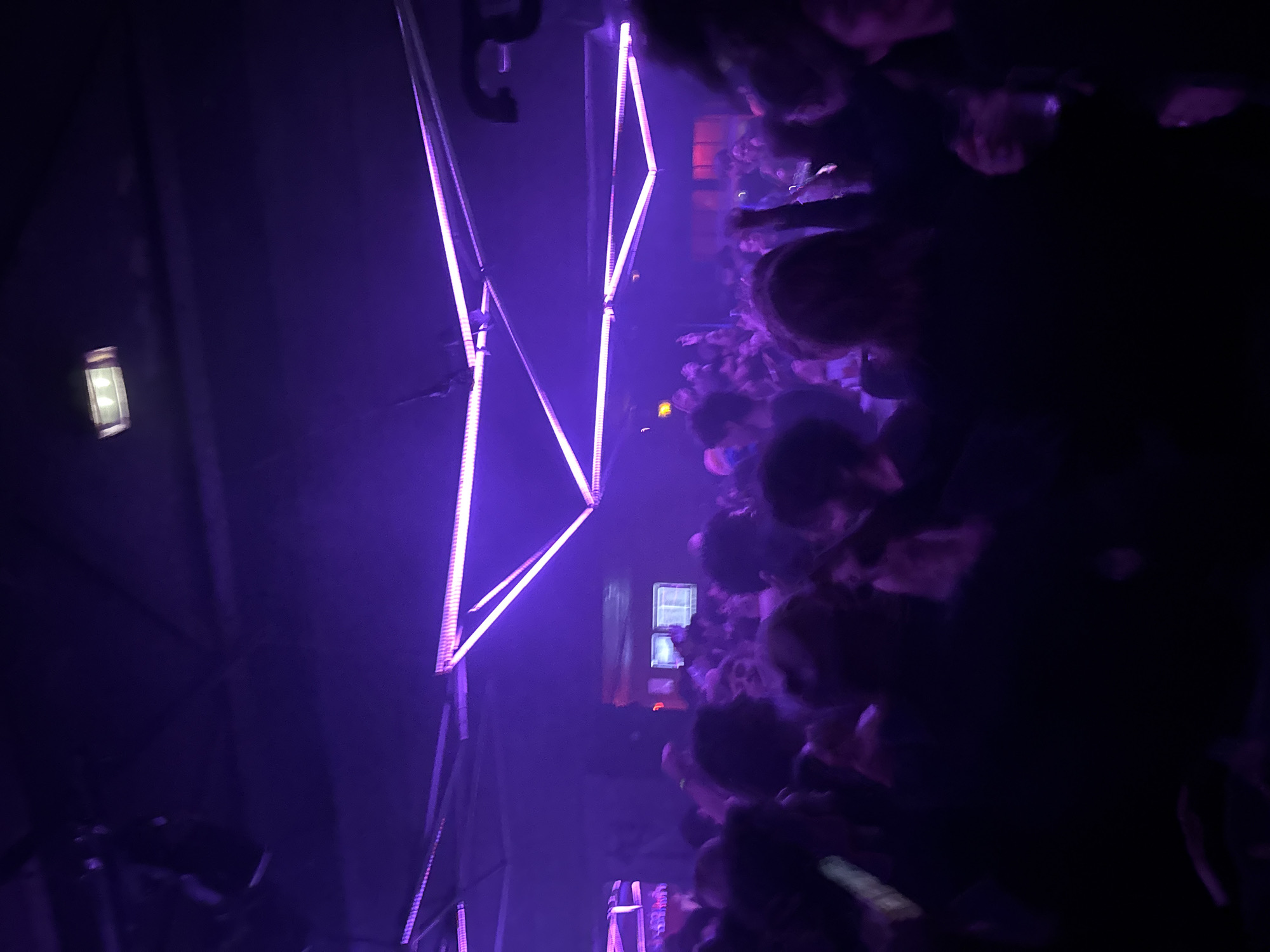 The image displays a densely packed indoor scene, consistent with a nightclub or music venue, characterized by low light and a dominant purple illumination. The ceiling features an elaborate lighting rig composed of linear LED strips arranged in geometric patterns, including prominent triangles and parallel lines emitting a bright purple glow. A rectangular light fixture is visible on the ceiling. A large crowd of individuals occupies the entire frame, seen primarily from behind or in profile. Their features are largely indistinct due to the low lighting and movement. People appear to be standing, socializing, and some have hands raised, indicative of dancing or engagement with an event. In the background, areas of red light are visible, suggesting additional lighting or features within the venue. Some individuals in the foreground are holding clear cups. The atmosphere appears hazy. This scene is situated within a commercial establishment in Paris, France. The specific location within the city is not visually discernible.
