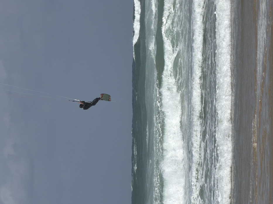 The image depicts a kitesurfer in mid-air over the Mediterranean Sea, off the coast of Tel-Aviv, Israel. A single individual, dressed in a dark full-body wetsuit and a dark helmet, is suspended several meters above the water, holding a light-colored control bar with both hands. Four thin white lines extend upwards from the control bar, indicating an unseen kite. A green and red/orange kitesurfing board is attached to the individual's feet, angled downwards, with water droplets visible beneath it. The sea is characterized by multiple parallel lines of breaking waves with white foam, extending across the frame from mid-ground to the foreground. The water appears choppy and a deep teal-green. The immediate foreground shows a strip of wet, light brown sand. The sky above is overcast, a uniform dark grey-blue, occupying the upper two-thirds of the frame. The scene captures an active watersport moment under cloudy conditions.