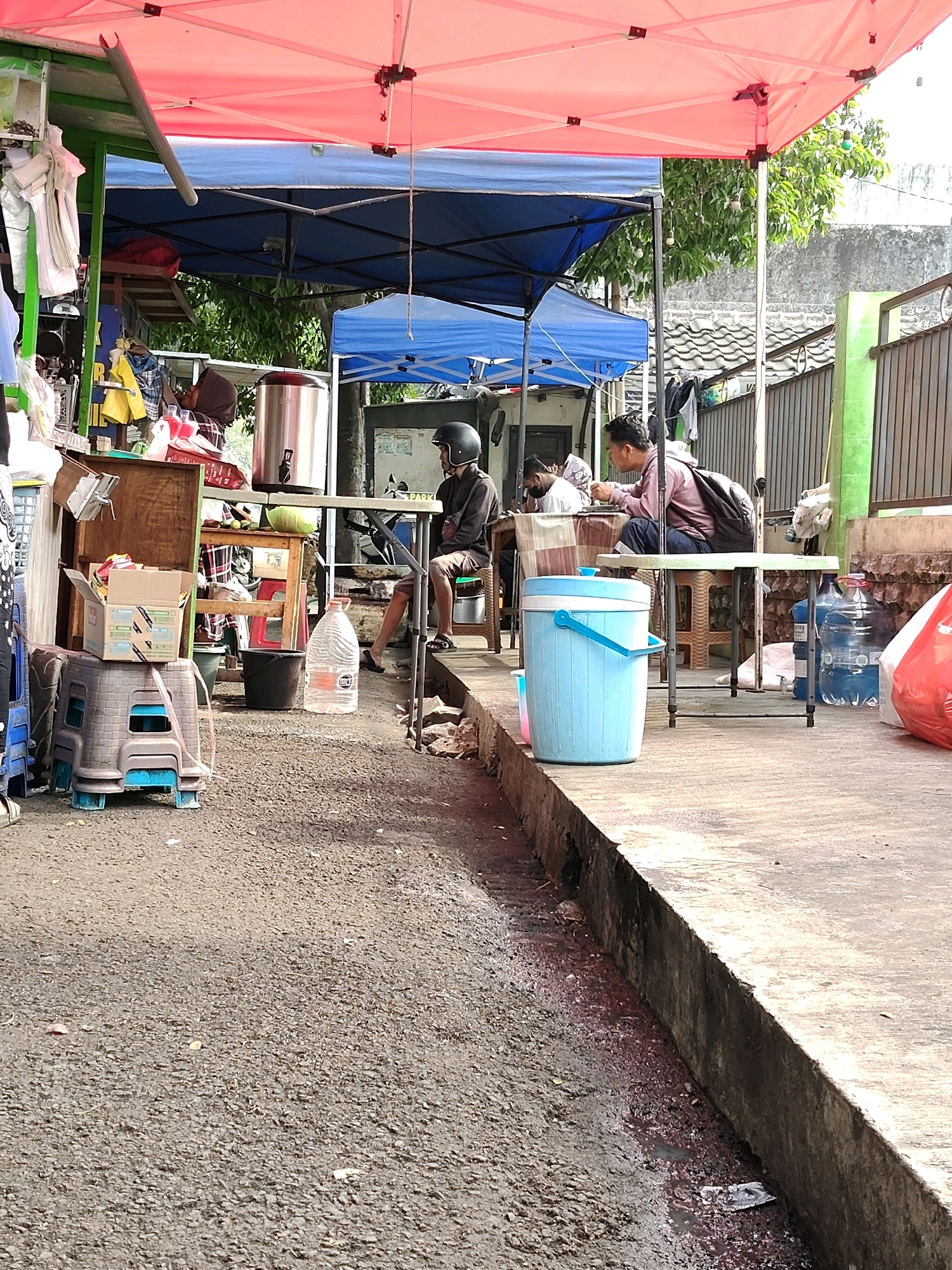 The image depicts an outdoor scene, likely a street-side market or food stall, in the city of Malang, Indonesia. In the foreground, a textured asphalt surface leads to a concrete curb and walkway. A large, light blue plastic cooler with a white lid and handle sits on the concrete walkway. Several large blue plastic water bottles are visible behind the cooler, with one large clear plastic bottle with an orange cap positioned to the left.
Behind the cooler and water bottles, a small group of people are seated at tables under a pink and blue canopy. Two men are the primary subjects. One man, wearing a dark jacket and a helmet, is seated on a stool, facing away from the camera. Another man, wearing a light pink shirt and carrying a backpack, is seated at a table, looking to his right, with his hands in front of him. A third person, partially visible, appears to be wearing a headscarf and is seated at the same table.
Various items are scattered around the scene, including a wooden crate with boxes on top, a black bucket, and what appears to be a large metal beverage dispenser. The backdrop includes a building with a tiled roof, a metal fence, and trees. The overall impression is one of a busy, everyday scene in a local commercial area.