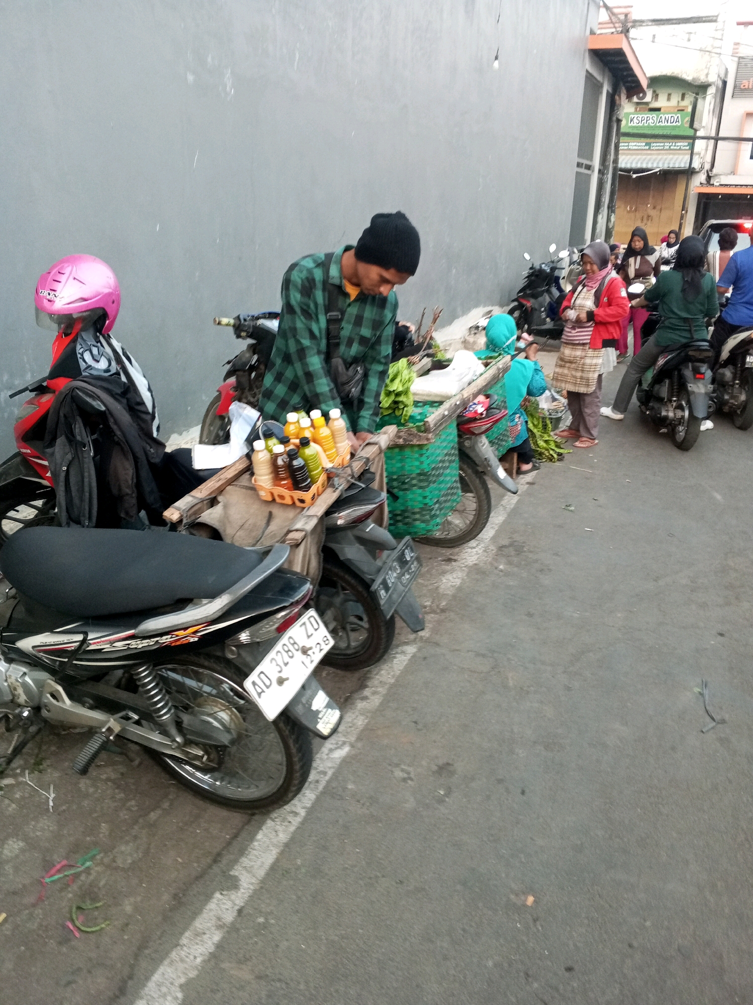 The image depicts a street scene in Gentan, Indonesia, with a focus on a roadside vendor and surrounding activity. The foreground shows the rear of a motorcycle with license plate AD 3288 ZD. To the left, a pink helmet rests on a motorcycle. A vendor, wearing a black beanie and a green and black plaid shirt, is bent over a cart loaded with bottles of beverages. The cart is green and woven, with a wooden frame. Several people, including women wearing headscarves, are visible in the background, some standing near motorcycles and others interacting with the vendor. The environment appears to be a street or sidewalk alongside a building with some signage, including "KSPPS ANDA". The overall scene captures a moment of daily commerce in an urban Indonesian setting.