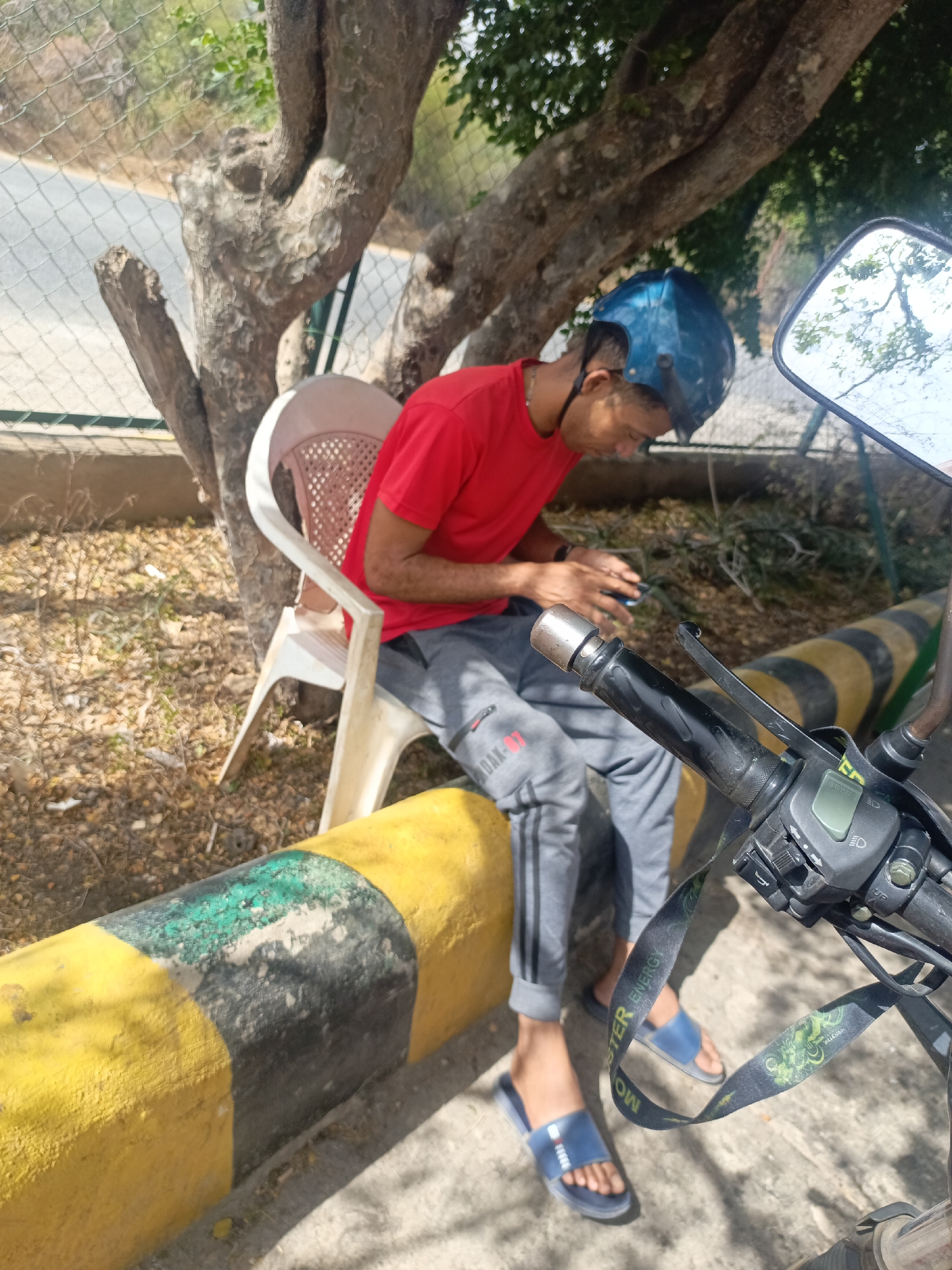 A young man in a red shirt and gray sweatpants is seated on a white plastic chair beside a tree, adjacent to a yellow and black striped concrete barrier in La Tagua, Venezuela. He is wearing a blue helmet and blue sandals, and is looking at a cell phone in his hands. The handlebars of a motorcycle, including the mirror and a lanyard with the words "MONSTER ENERGY" on it, are visible in the foreground. Behind the tree is a wire fence. The scene is outdoors, in daylight.
