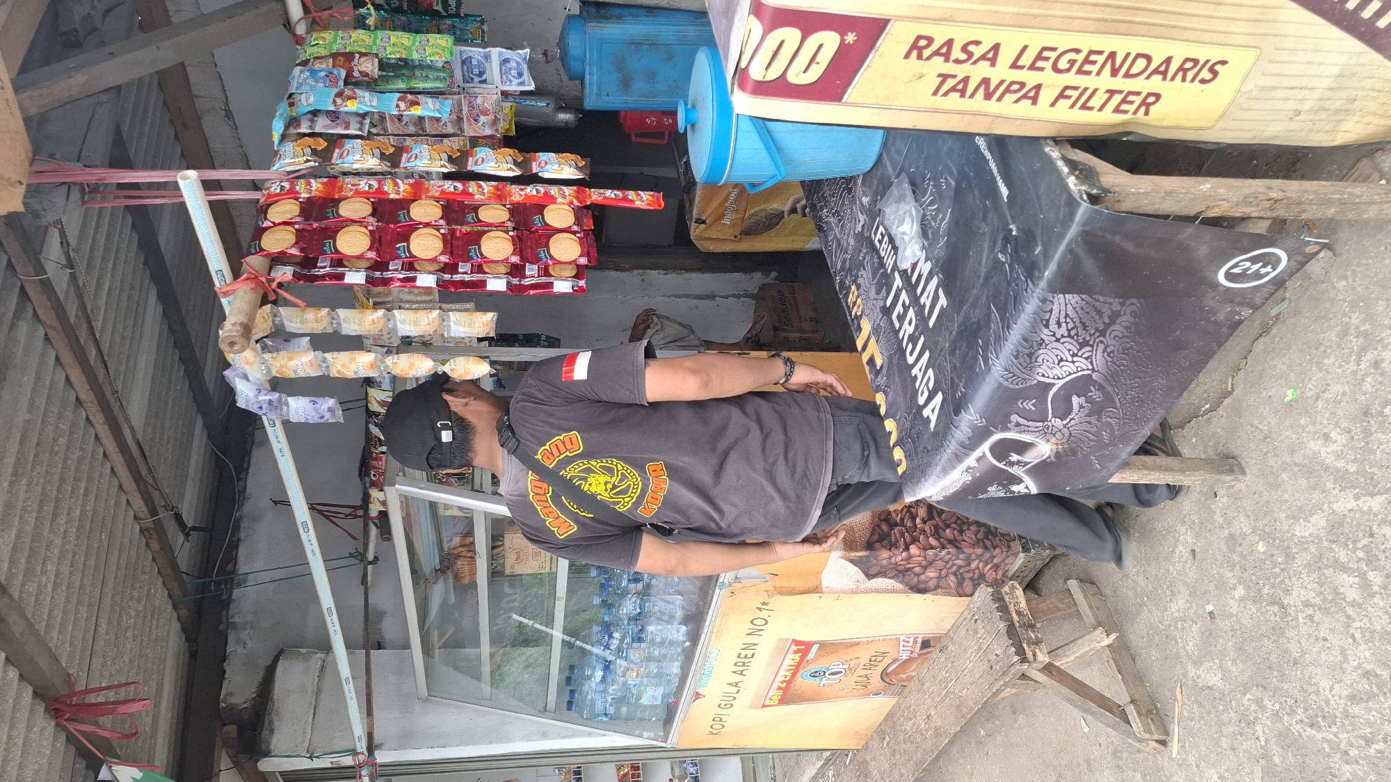 A middle-aged Indonesian man wearing a black baseball cap and a dark t-shirt with a yellow and black emblem and an Indonesian flag patch on his sleeve stands facing away from the camera at a small roadside stall. The stall is open-air, with a corrugated metal roof supported by wooden beams. Shelves made of bamboo and pipes hold a variety of packaged snacks and drinks, including bottled water in a refrigerated display case. A wooden bench is positioned to the left of the stall, and a table covered with a black tablecloth featuring an ornate pattern stands in front of it. The man appears to be browsing the goods. The ground is paved with concrete. In the background, a large blue cooler and a red container are visible. The scene suggests a typical small business operating in a community setting in Cilaku, Indonesia.
