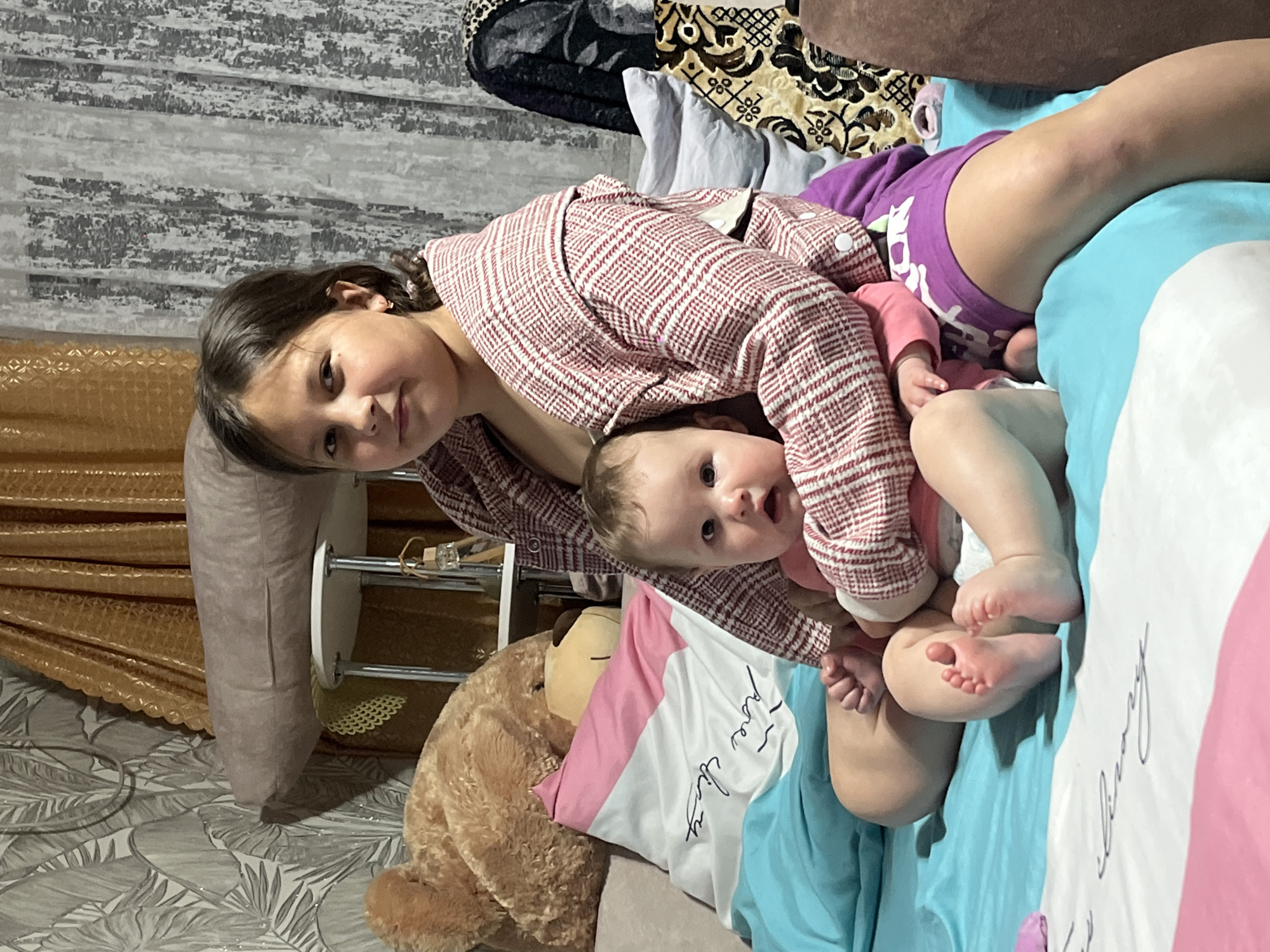 Two children are on a bed. One child, an older girl with brown hair, is wearing a red and white patterned robe and purple shorts. She is leaning over a baby. The baby, who is wearing a pink outfit, is lying on the bed. The bed has light blue and white bedding with pink accents. A large teddy bear is positioned next to the bed. A side table with a golden-colored curtain behind it is in the background. The floor is covered in a gray and white patterned material. The setting is indoors, and the location is specified as Novopokrovskaya, Russia.