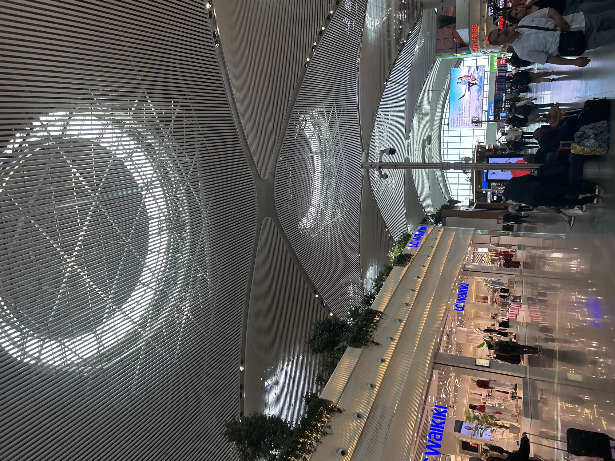The image captures the interior of a vast, modern airport terminal, likely Istanbul Airport in Arnavutköy, Turkey, given the architectural style and visible brands. The scene is oriented as if the camera was rotated, making the floor appear on the right and the ceiling on the left.
The most striking feature is the intricate ceiling design, which spans the upper portion of the image. It consists of a wave-like structure made from numerous horizontal slats and curved, perforated panels, creating a sense of depth and movement. Bright, diffused light emanates from behind these structures, casting reflections on the polished floor below.
Numerous people, presumably travelers, populate the terminal. They are scattered throughout, some standing, others seated in waiting areas. Many have luggage near them, indicative of their travel purpose. While individuals are not clearly distinguishable, their presence contributes to the bustling, active atmosphere of a major transportation hub.
On the right side of the image, retail establishments line a corridor. A prominent storefront for "LC Waikiki" is clearly visible, displaying mannequins dressed in clothing. Other blurred shopfronts and large digital advertising screens are also present. One screen in the background features an image of an airplane and people, with text partially visible, including "UNBELIEVABLE FREE" and "SHOP&FLY", and "DUTY FREE" on another sign above a kiosk. A large vertical pillar in the mid-ground supports a surveillance camera, and decorative greenery lines some of the architectural elements.
The lighting suggests an indoor setting, possibly evening or night, with strong artificial illumination. The overall impression is one of a spacious, well-designed, and busy travel environment.