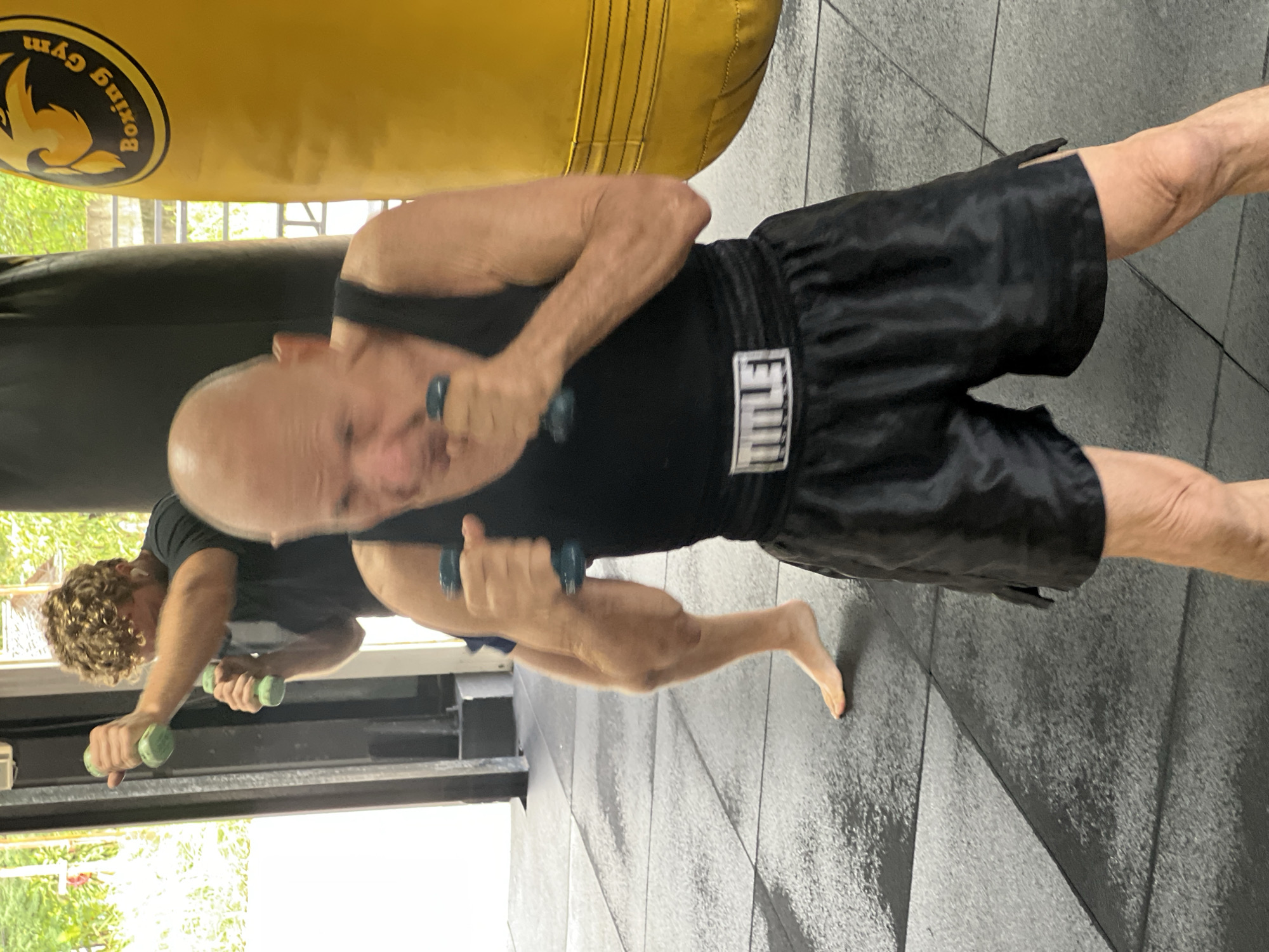 The image depicts a boxing gym in Chalong, Thailand, as evidenced by the "Boxing Gym" logo visible on a yellow punching bag in the background. Two individuals are engaged in a boxing or fitness training session. In the foreground, a bald, older man wearing a black tank top and black boxing shorts with a white "TITLE" logo on the waistband is holding light blue dumbbells in each hand, positioned as if preparing to punch. His posture suggests an active stance. In the middle ground, another man with curly blond hair, wearing a dark t-shirt, is also holding two green dumbbells, his arms bent and raised, indicating a similar training exercise. The gym floor is a black and gray tiled surface, with some areas appearing wet, possibly from sweat or cleaning. The environment includes gym equipment such as punching bags and natural light filtering in from the background.