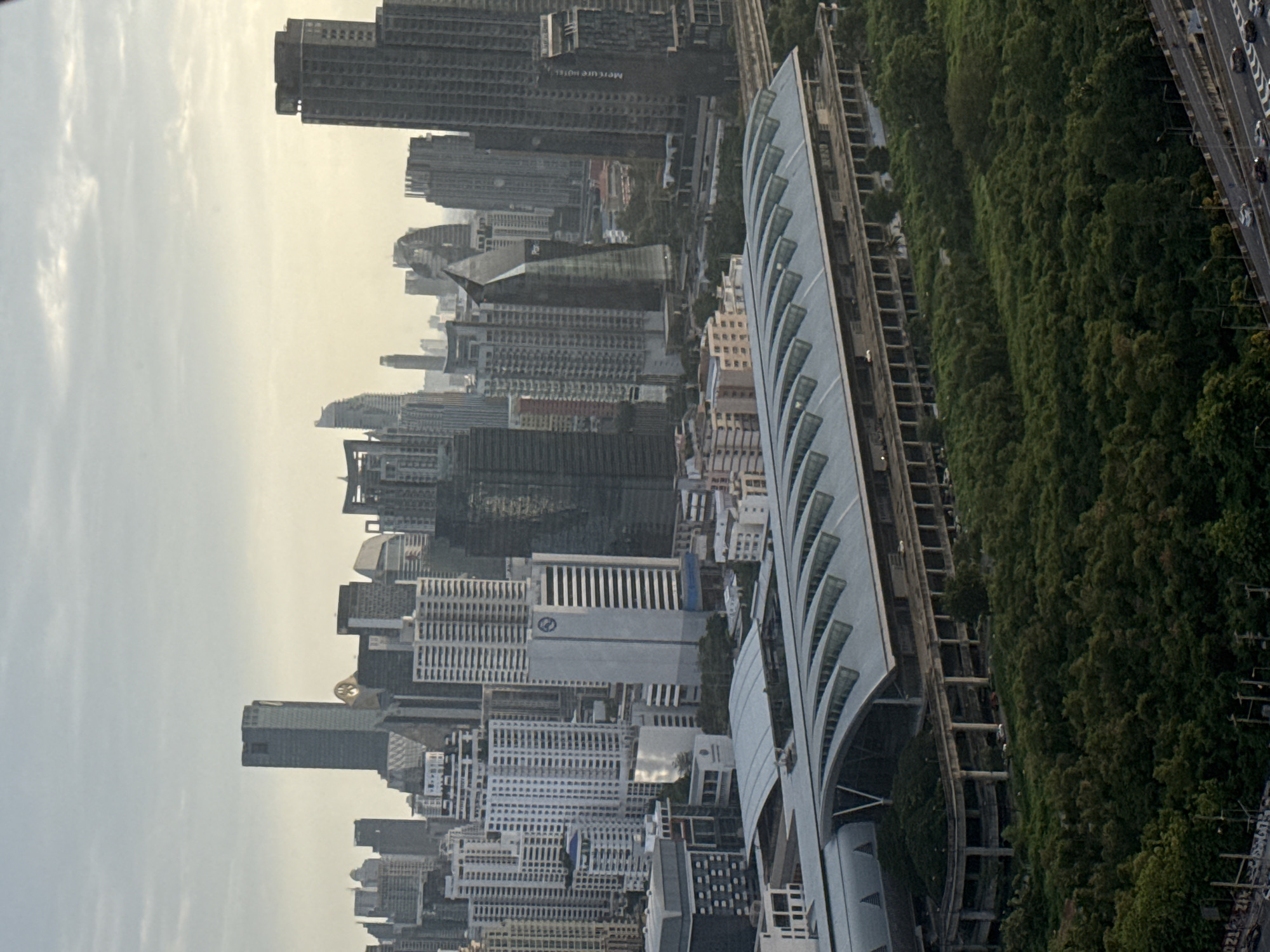 This is an aerial, slightly angled, vertical shot of a city skyline in Bangkok, Thailand. The image captures a large, modern train station with a distinctive curved, silver roof dominating the lower half of the frame. A dense canopy of lush green trees surrounds the station. Behind the station, a densely packed cityscape of numerous skyscrapers of varying heights and architectural styles rises into a hazy, overcast sky. The buildings are a mix of glass, concrete, and metal. The time of day appears to be late afternoon or early evening, given the soft, diffused light and the muted colors. The overall impression is one of a vibrant, modern urban environment with elements of nature interspersed within it.