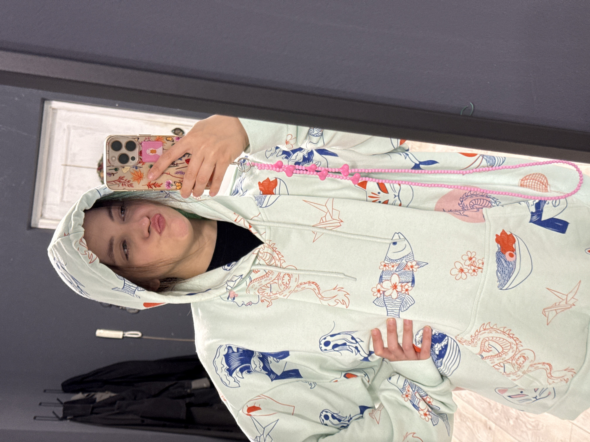 A young woman is taking a selfie in a mirror. She is wearing a light green hoodie with Japanese-themed designs, including fish, dragons, and origami birds. She is holding a phone with a floral case and a pink PopSocket. A pink beaded phone strap is draped around her neck. The background shows a dark wall, a window frame, and what appears to be a pile of black items, possibly jackets or umbrellas. There is no specific evidence in the image to confirm the location as Chaikovsky, Russia.
