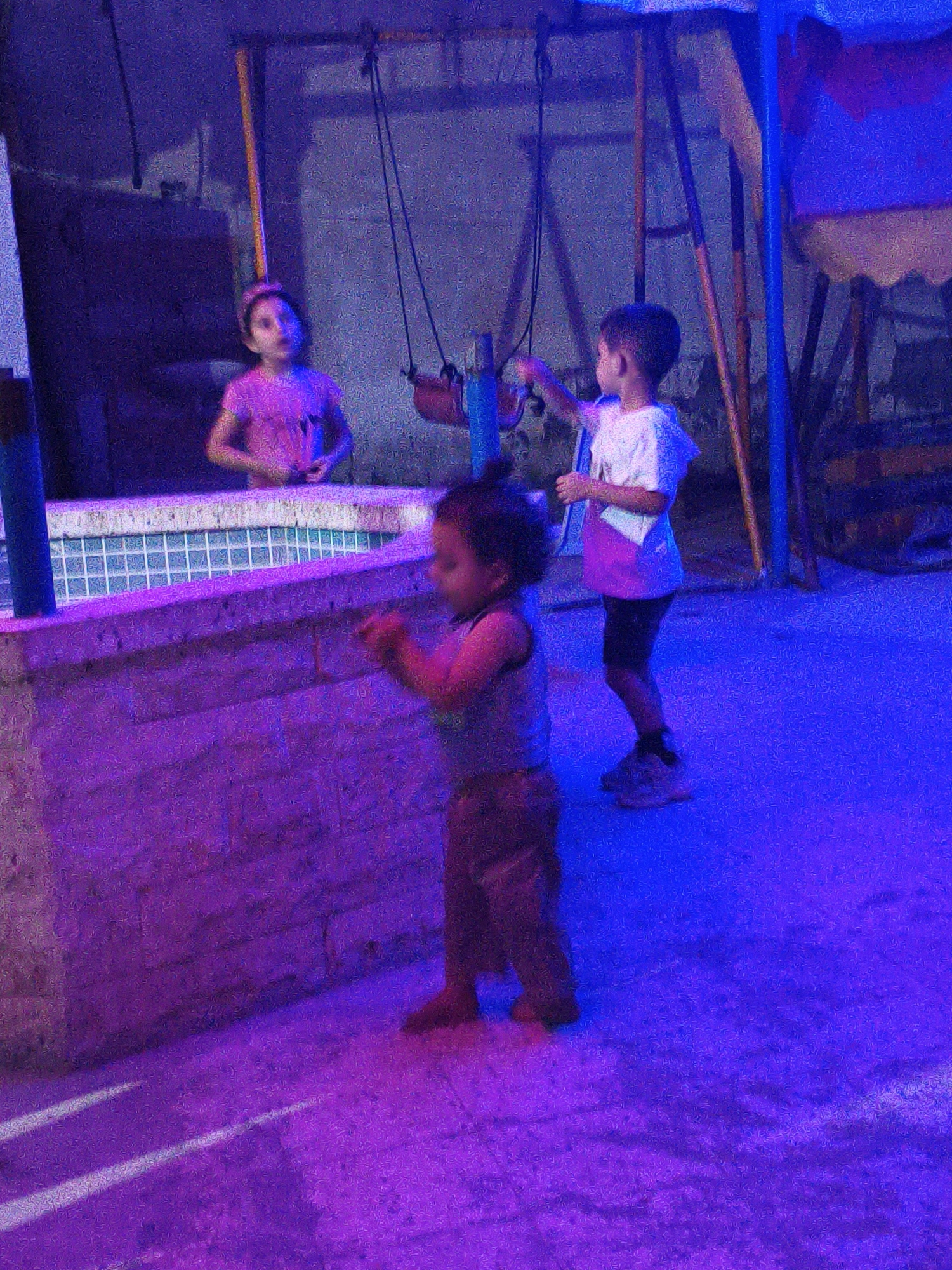 A medium shot captures three young children in what appears to be an outdoor play area with a swing set and a low wall. The scene is illuminated by a mix of purple and blue lights, creating a somewhat surreal atmosphere. The children are engaged in their own activities.
On the left, a young girl wearing a pink t-shirt stands near a tiled pool or water feature, her hands resting on a textured brick wall. Her gaze is directed away from the camera, and she appears to be looking at something out of frame.
In the center, a toddler wearing a light-colored sleeveless shirt and dark shorts stands on the ground, facing away from the camera. This child is holding something in their hands, though it is unclear what it is.
To the right, a young boy in a white t-shirt and dark shorts is standing. He is facing to the left and appears to be reaching towards the swing set structure with one hand.
The environment suggests an urban setting, possibly within a residential area or a public park. The presence of a swing set and what looks like a pool could indicate a space designed for children's recreation. The lighting suggests it might be evening or night, with artificial lights casting strong colors on the scene. The overall impression is one of ordinary childhood play, albeit under unusual lighting conditions.